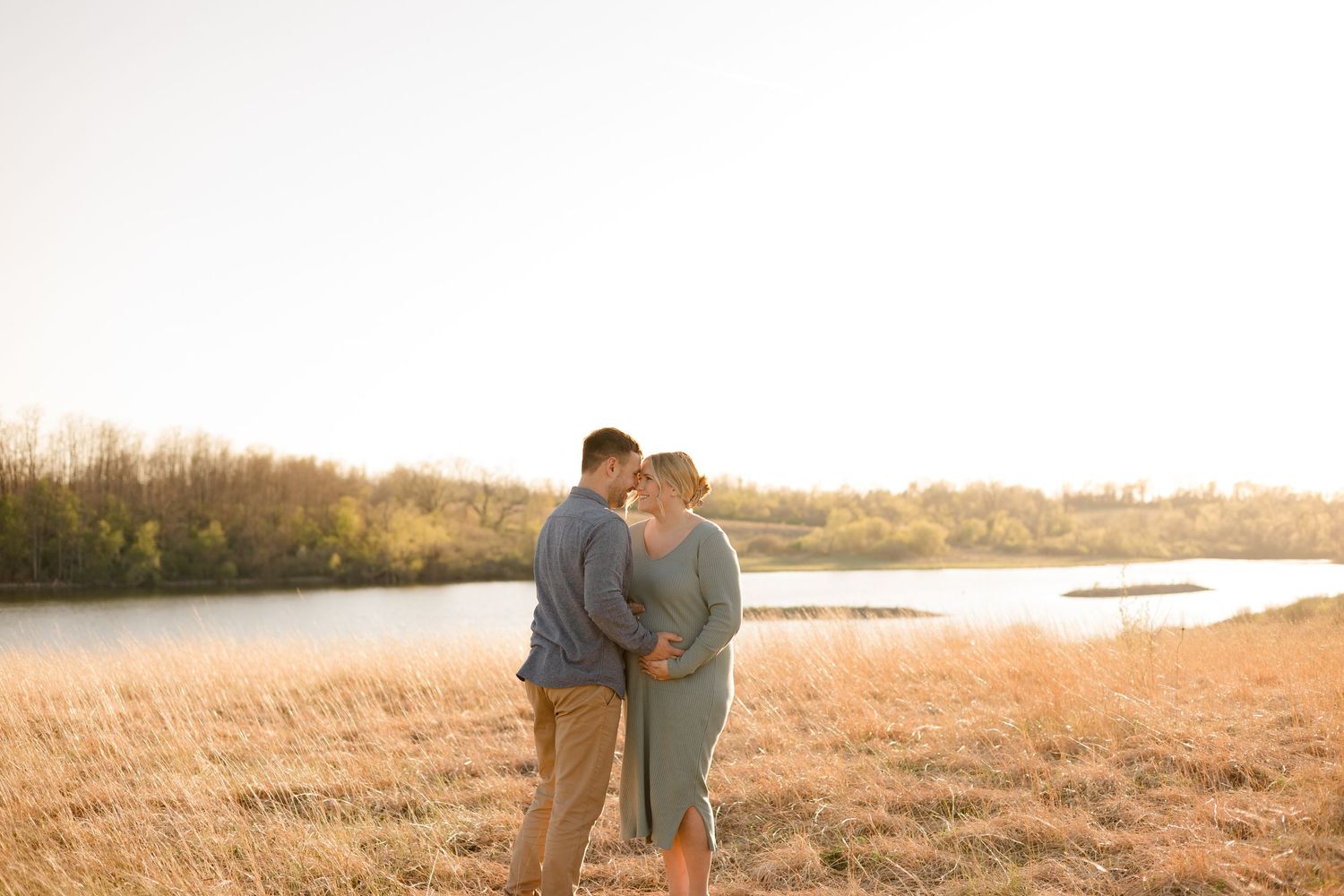 A couple stands close together in a golden field at sunset near a waterfront.