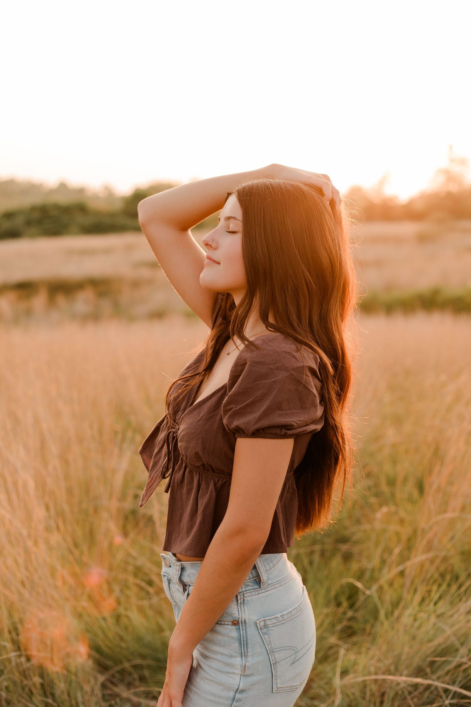Portrait of a young woman in brown top and denim against golden sunlit field at sunset.