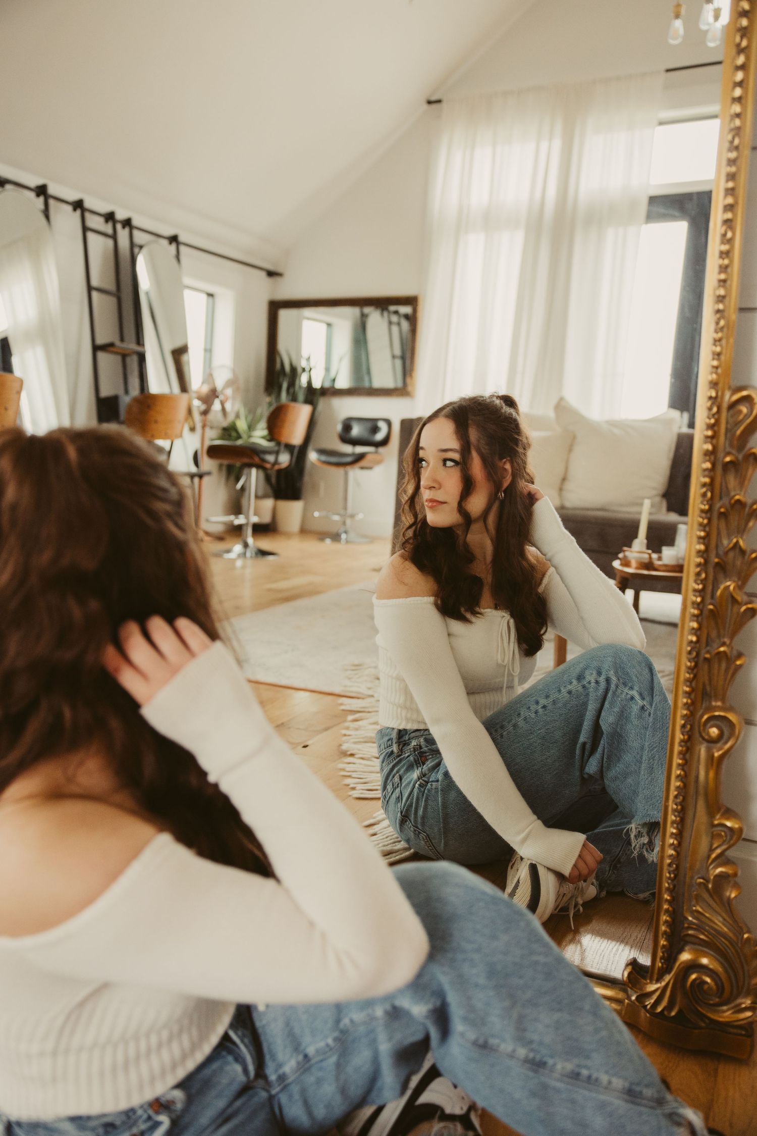Reflection in ornate gold mirror shows a high school senior in white sweater and jeans sitting on hardwood floor in sunny room.