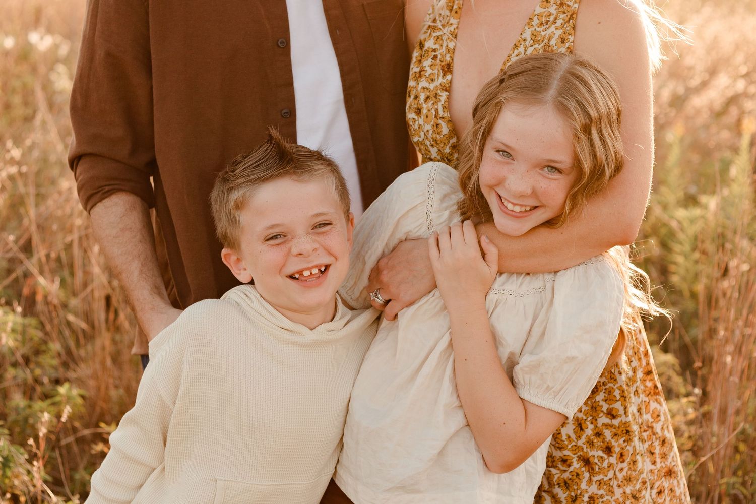 Golden outdoor family portrait in a field with warm sunlight illuminating joyful expressions and casual clothing.