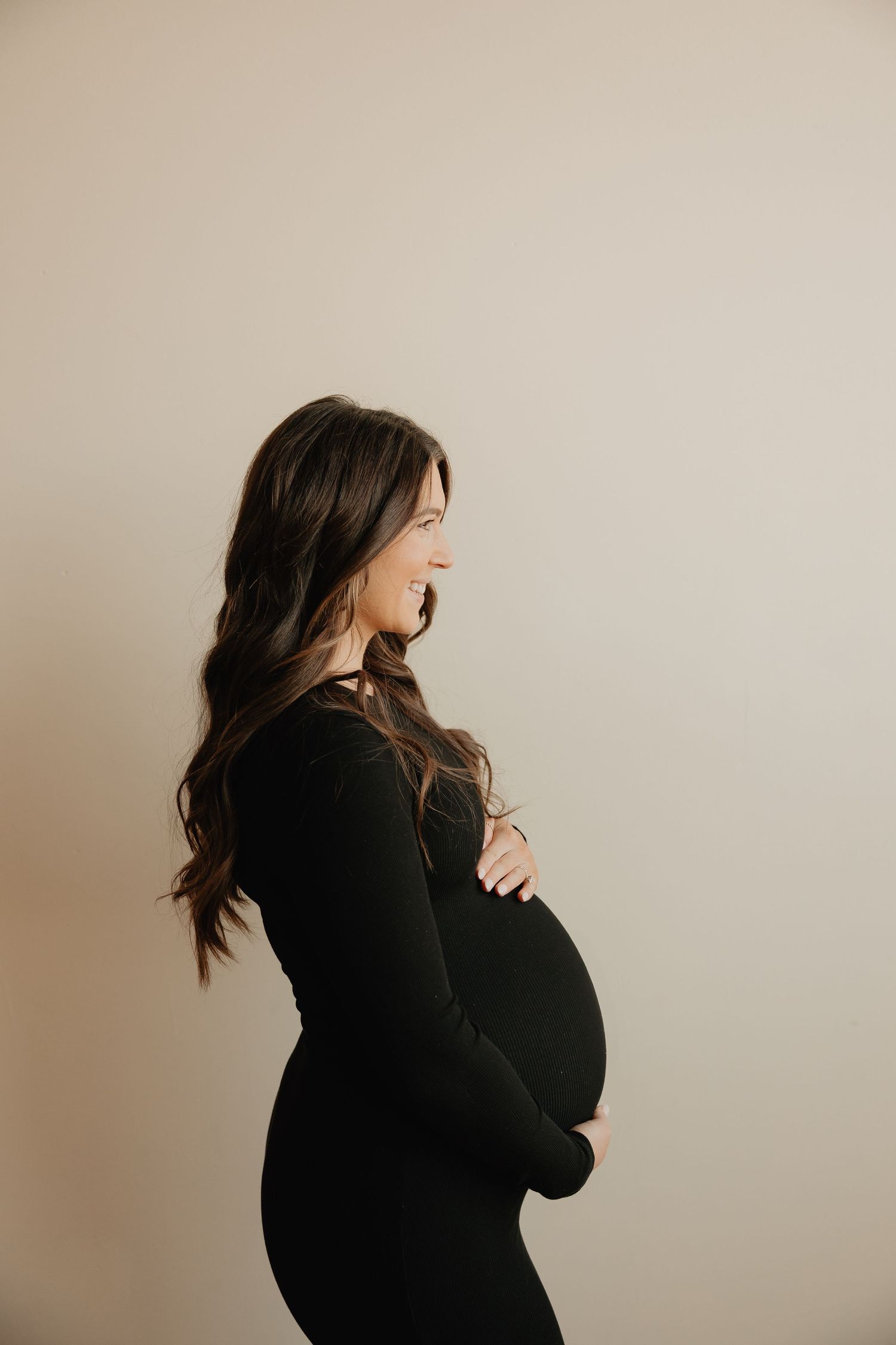 Side profile of person wearing black dress showing pregnant belly against beige background.