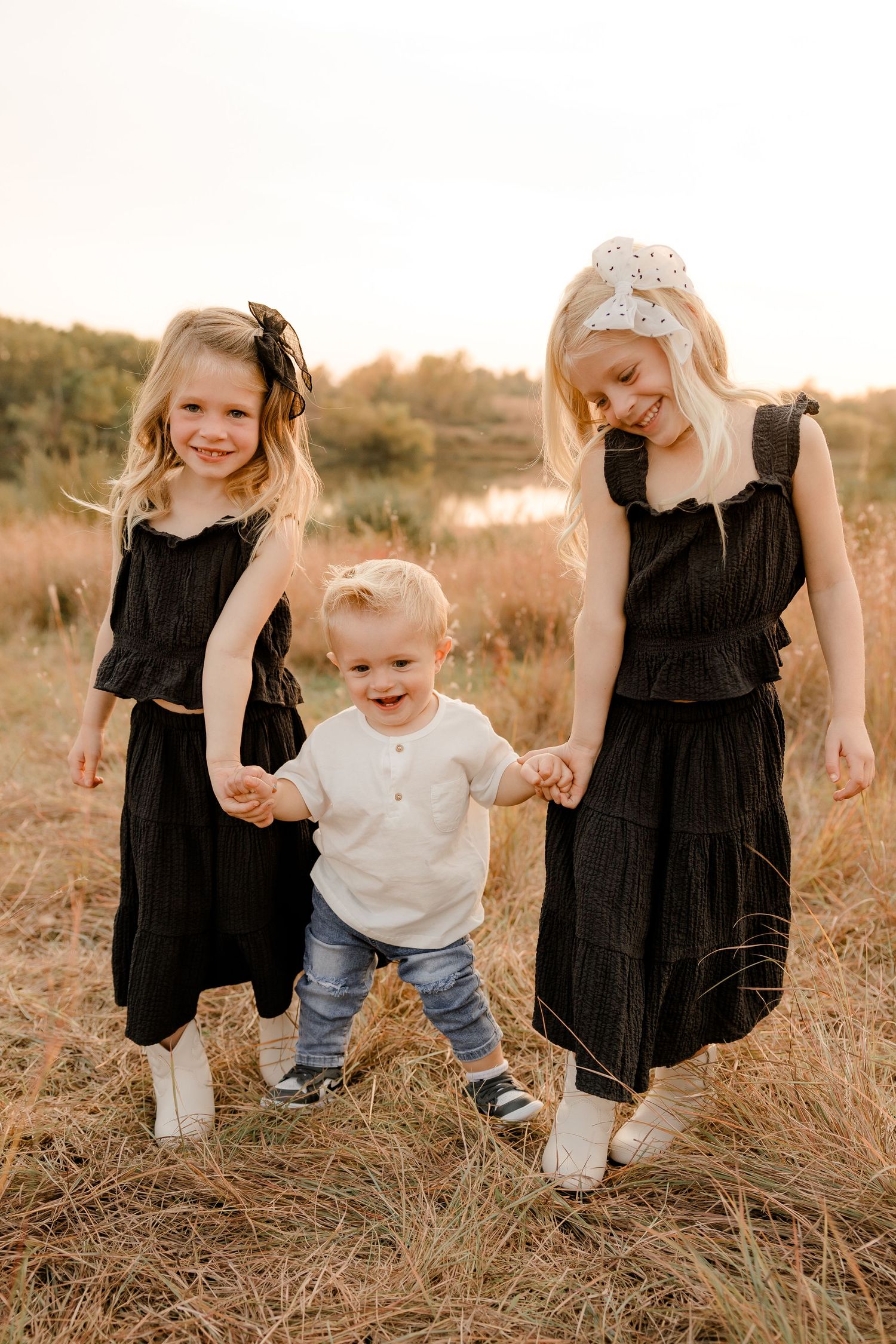 Three siblings hold hands and laugh together while walking in a golden field at sunset wearing black dresses and casual clothing.