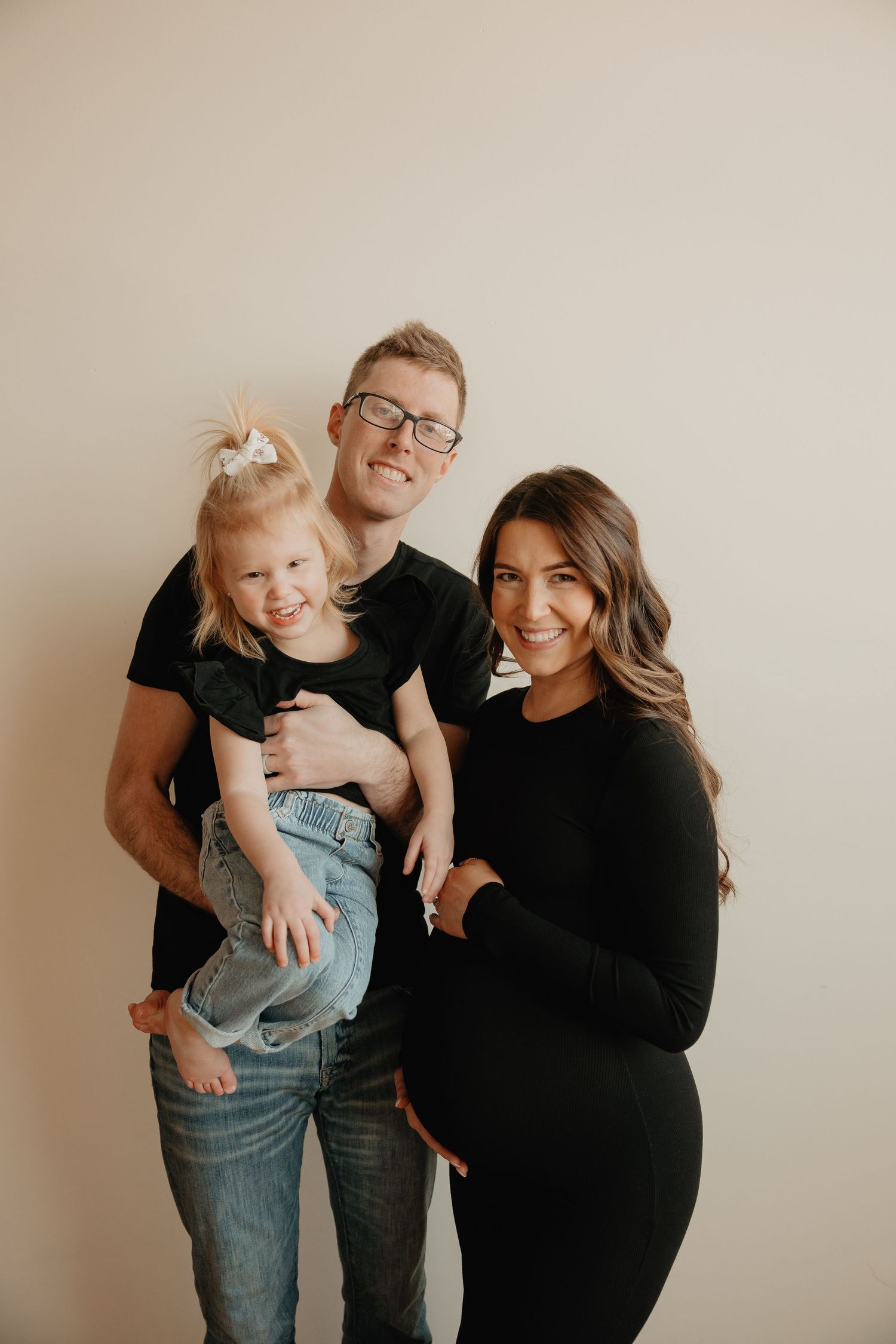 A group of three people wearing black tops pose together against a plain beige wall for a casual studio portrait.