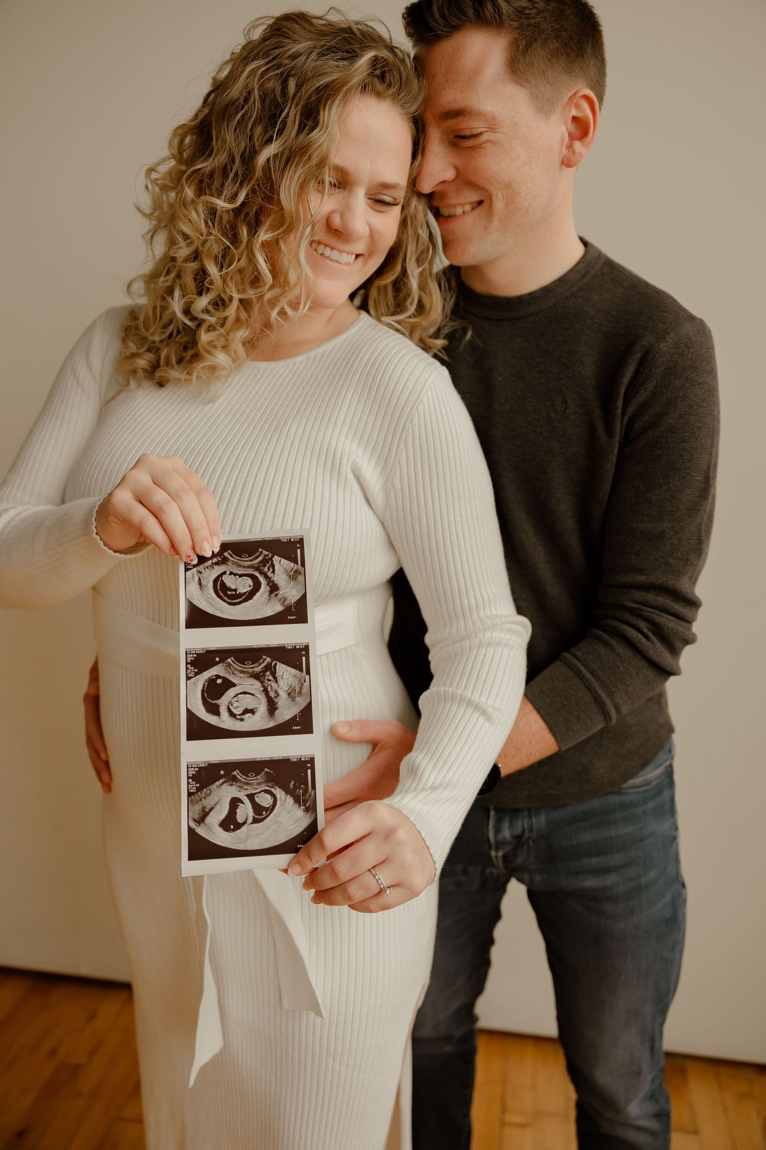 A couple shares an intimate moment while holding ultrasound photos of their expected baby in a natural light studio.