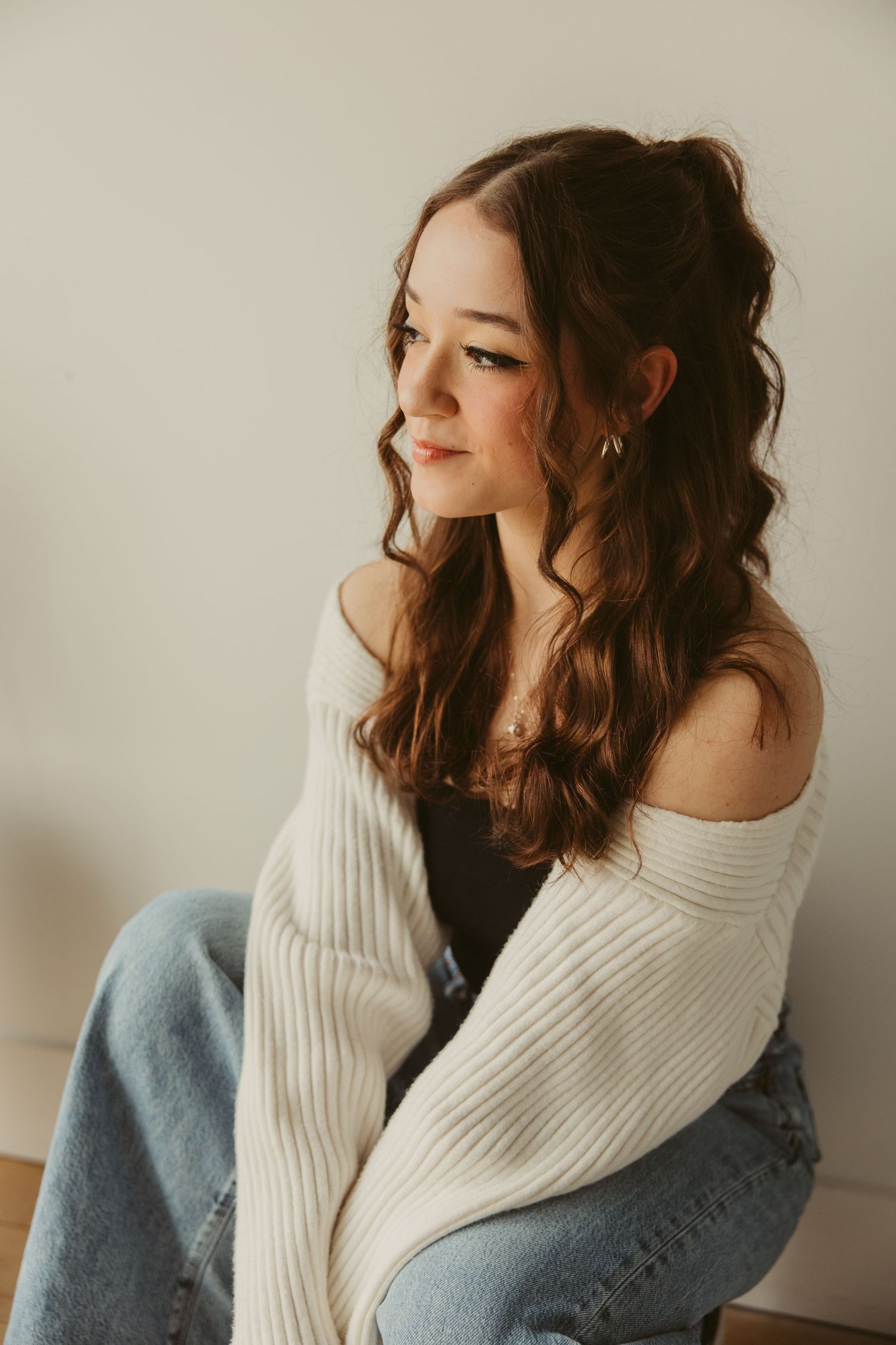 Portrait in white knit sweater and jeans poses against neutral wall in soft lighting with flowing brown hair.