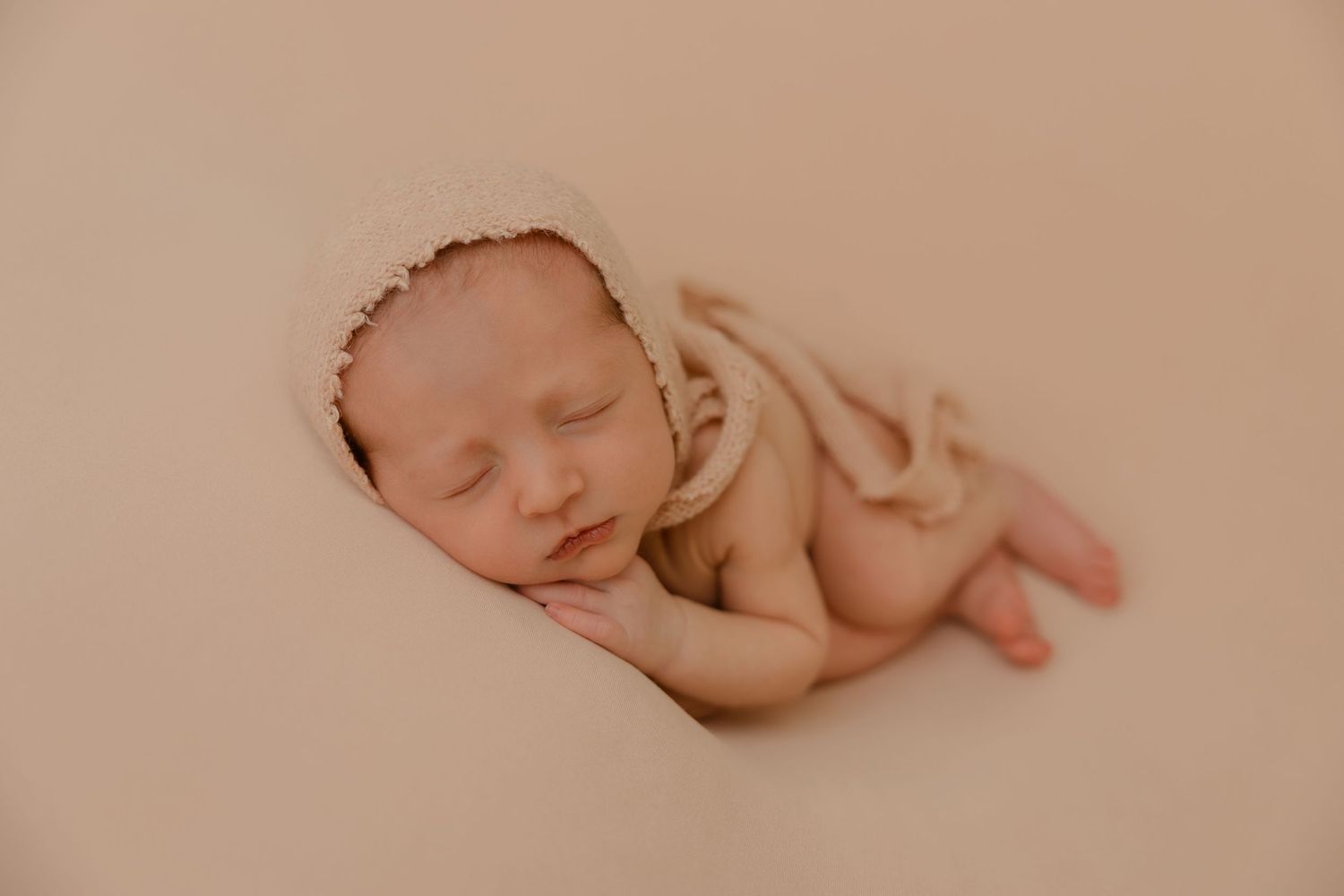 A peaceful sleeping newborn baby rests curled up on a soft beige backdrop in a gentle pose.