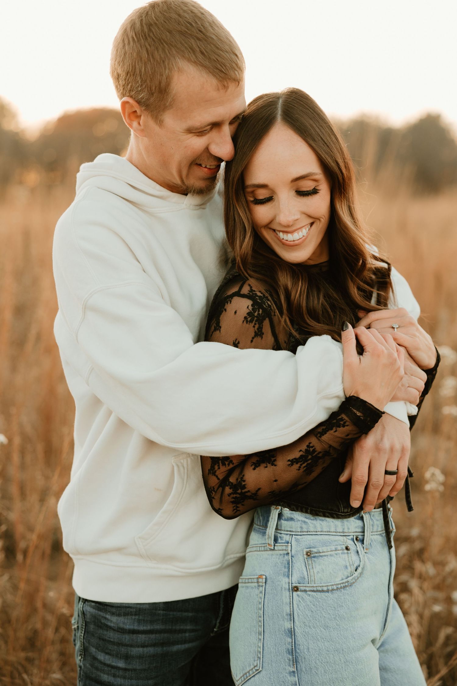 Romantic couple embraces in golden field at sunset while wearing casual white sweater and lace top.