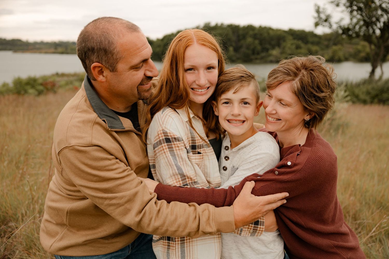 Family embraces together outdoors in a warm group hug against an autumn lakeside backdrop.
