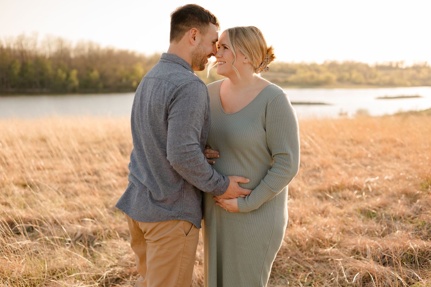 A couple shares an intimate moment in a golden field at sunset by the water's edge.