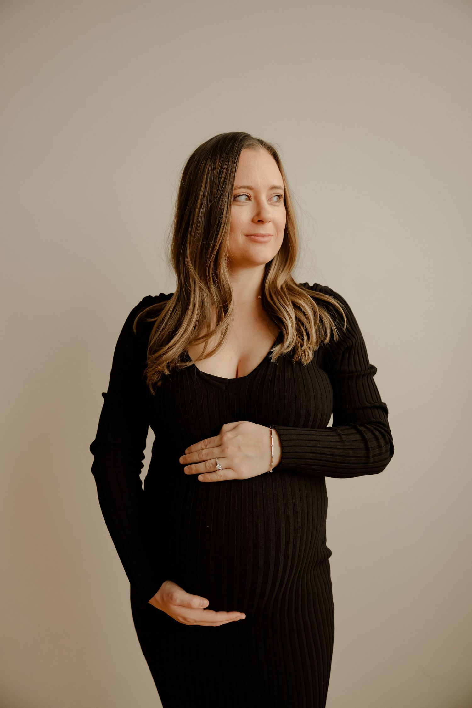 A pregnant woman in a black ribbed sweater dress poses against a beige background for a maternity photo.