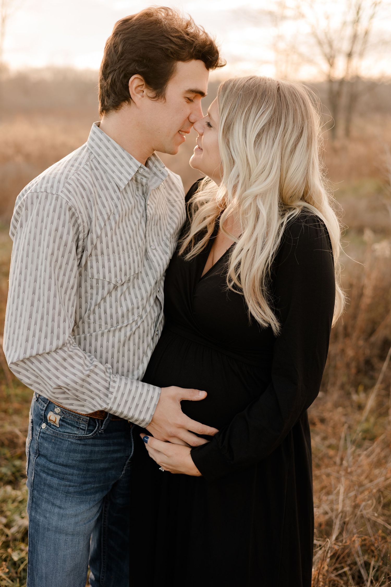 A couple shares an intimate moment in a field at golden hour, with her wearing black and him in a striped shirt and jeans.