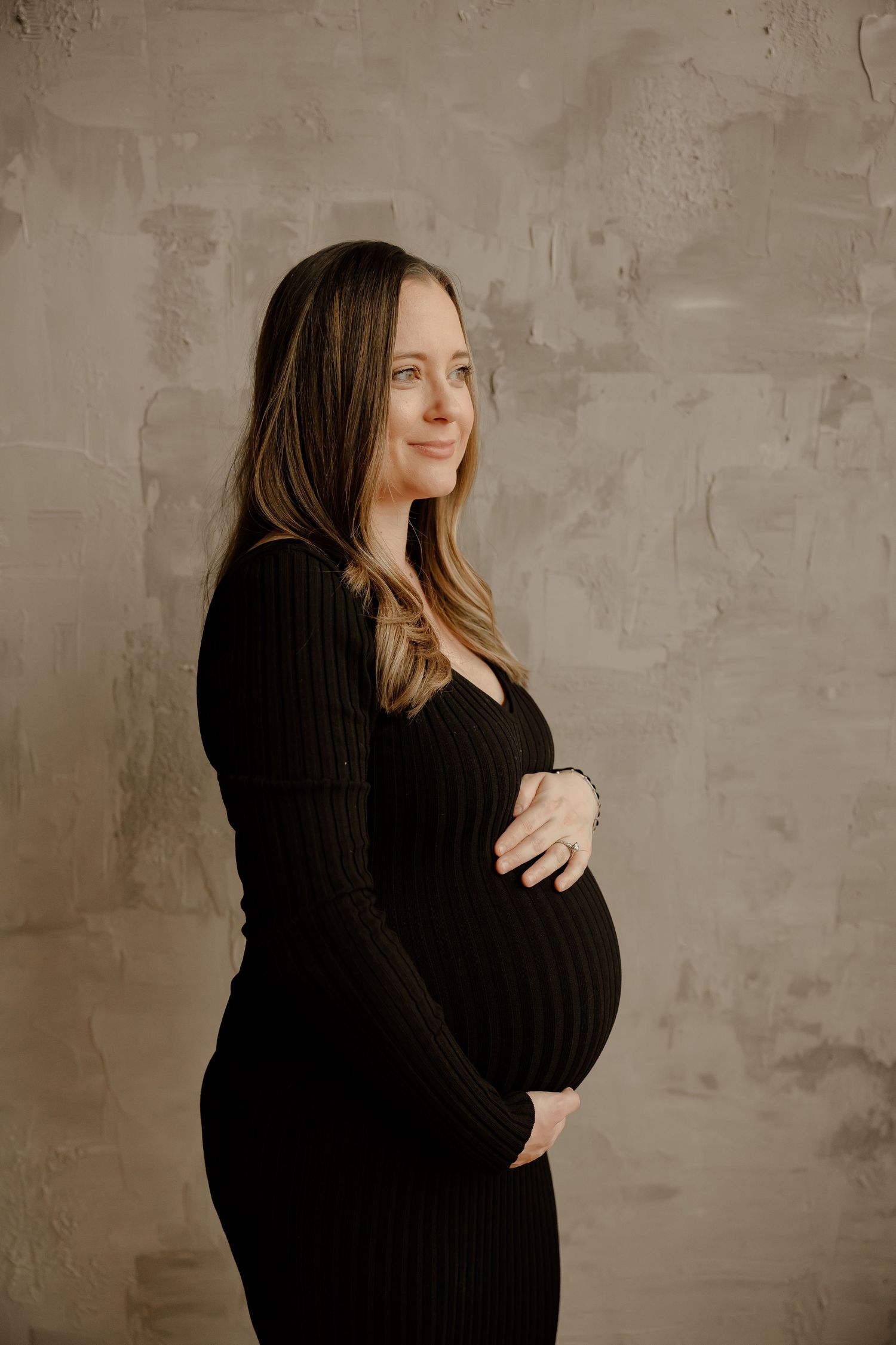 A pregnant woman in a black dress stands against a textured beige wall, gently cradling her belly.