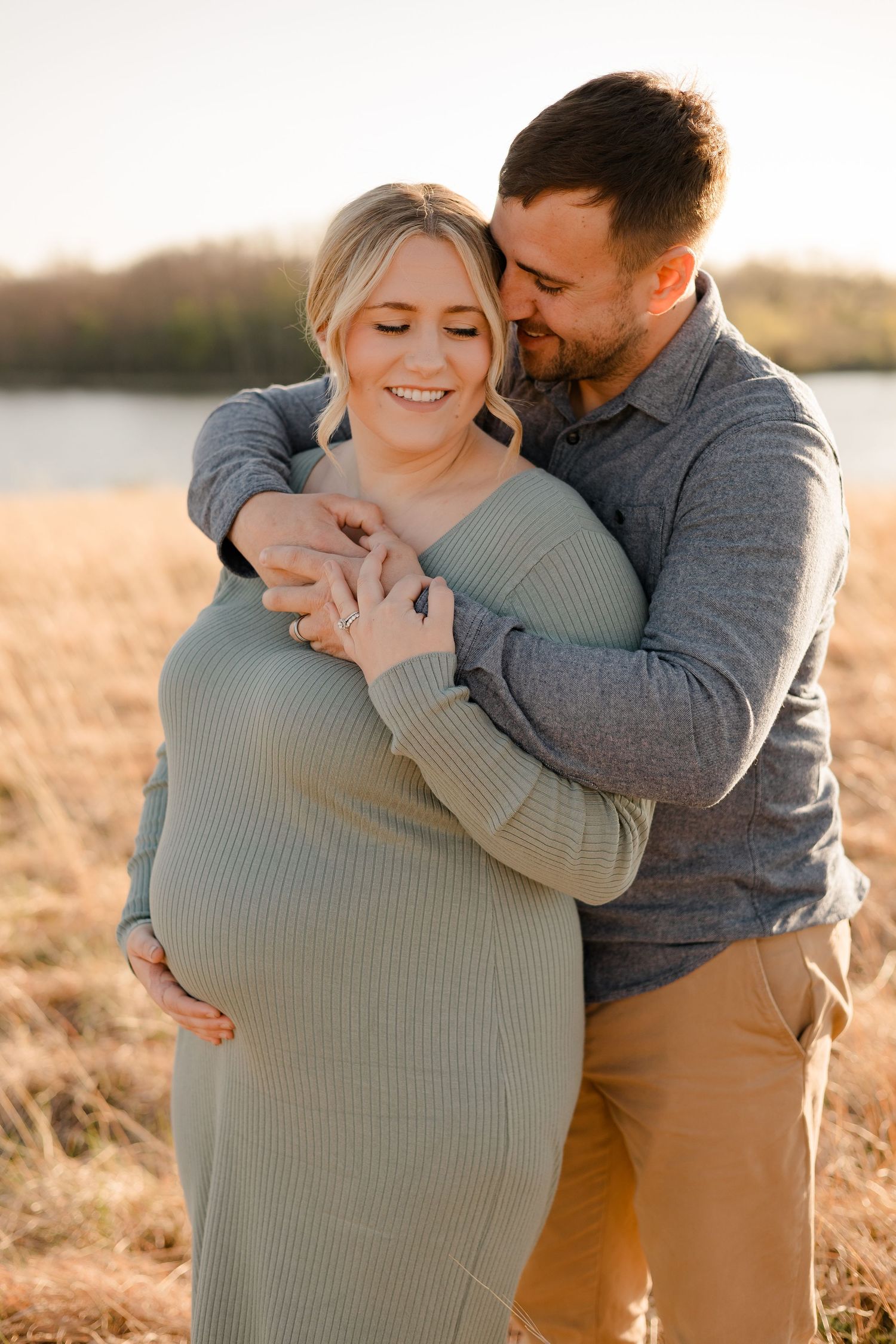 A couple embraces in a sunlit field during a maternity photo shoot at golden hour.