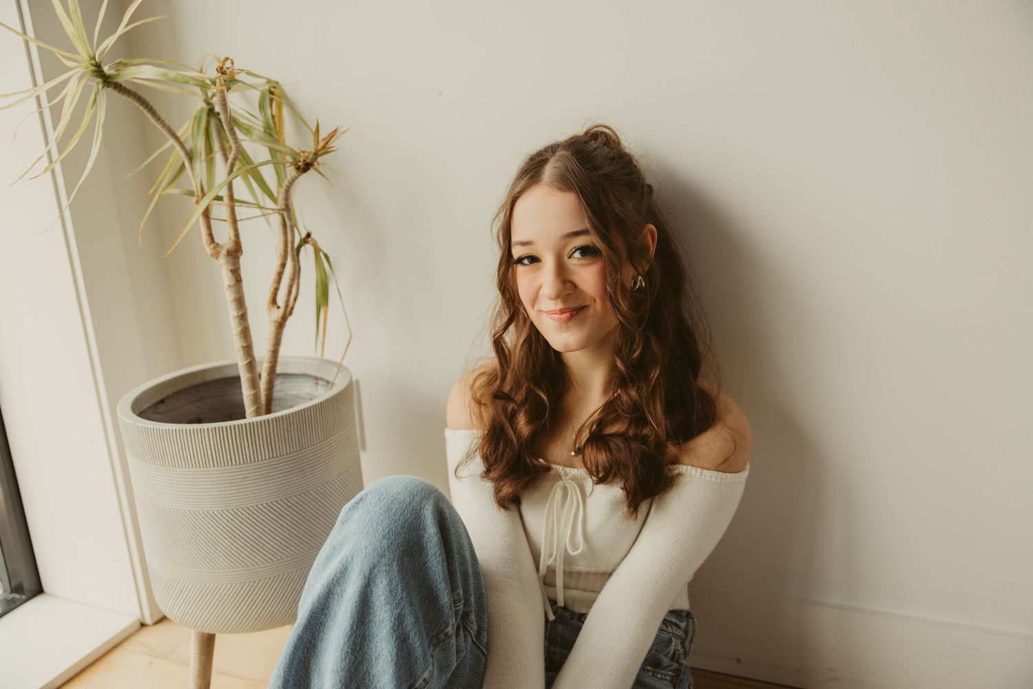 Lifestyle portrait of a high school senior with long wavy hair wearing an off-shoulder sweater sitting by window with houseplant.