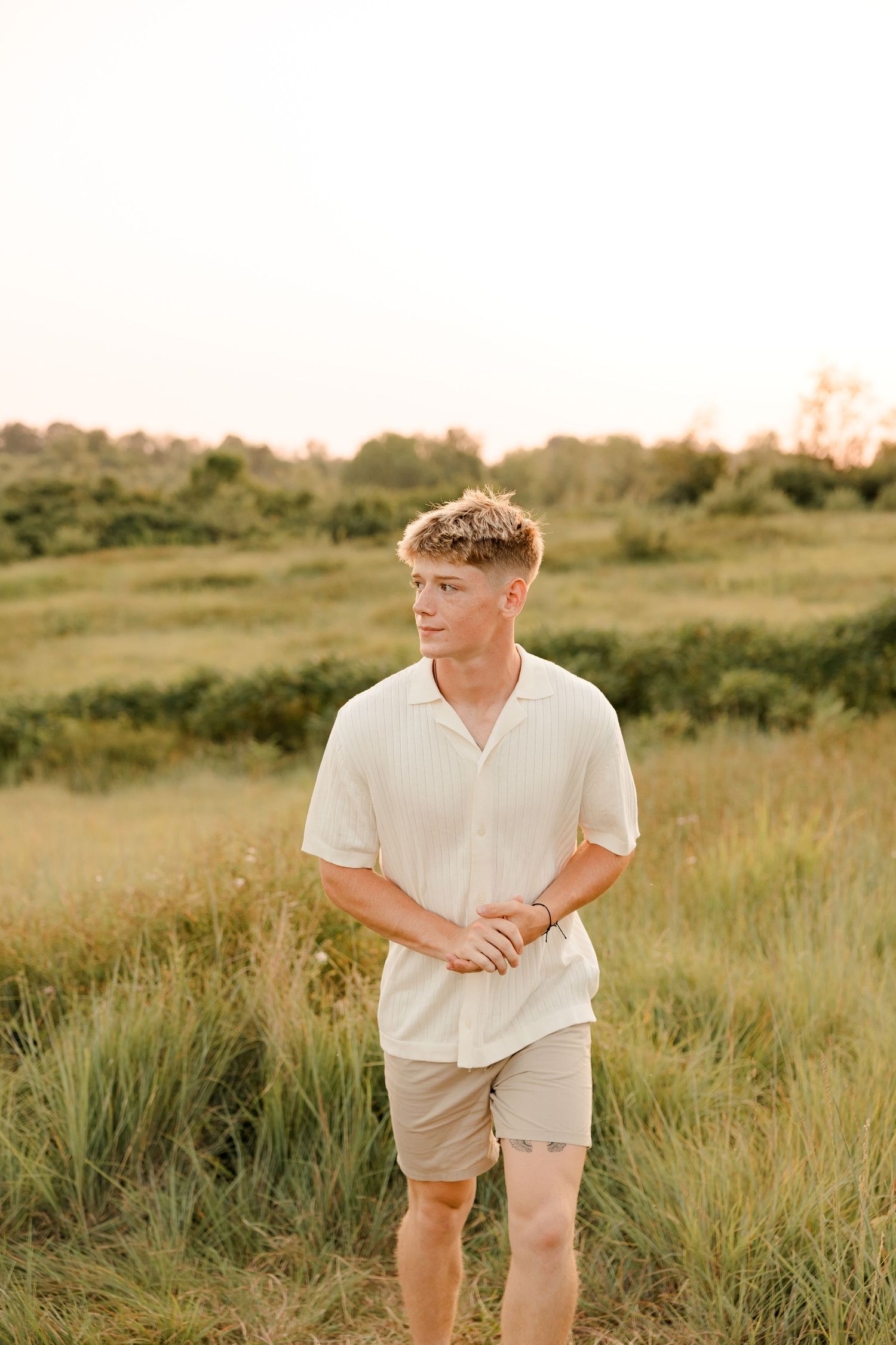 A high school senior stands in a golden field at sunset wearing a white polo shirt and khaki shorts against a rural landscape.