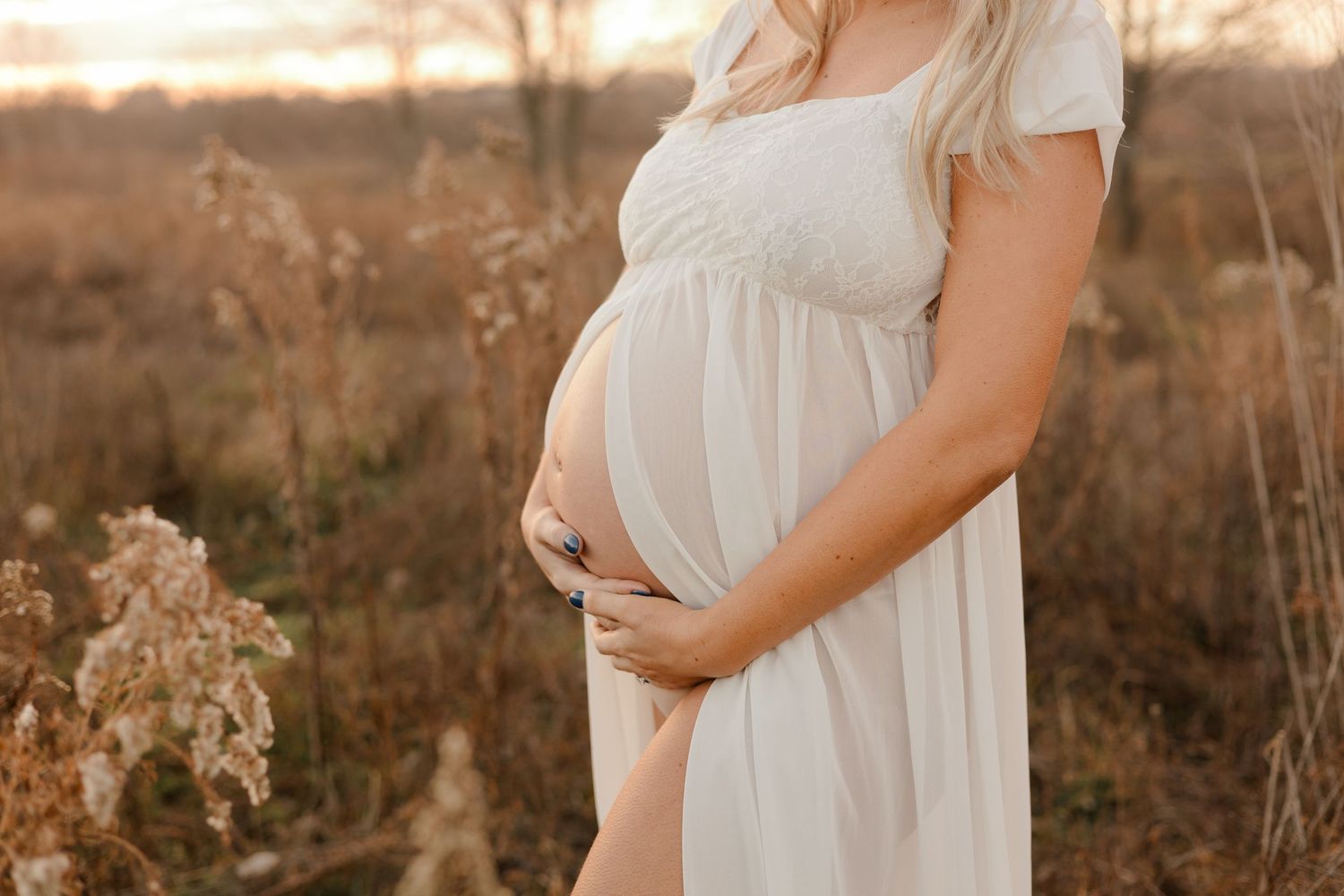 A pregnant woman in a flowing white dress cradles her belly during sunset in a rustic outdoor setting.