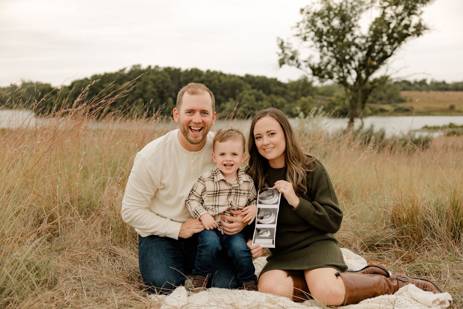 A family sits together on a blanket in a field of tall grass near a lake during golden hour.