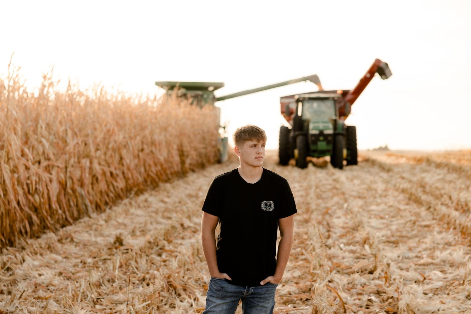A high school senior stands in a corn field at sunset while a combine harvester and tractor work in the background.