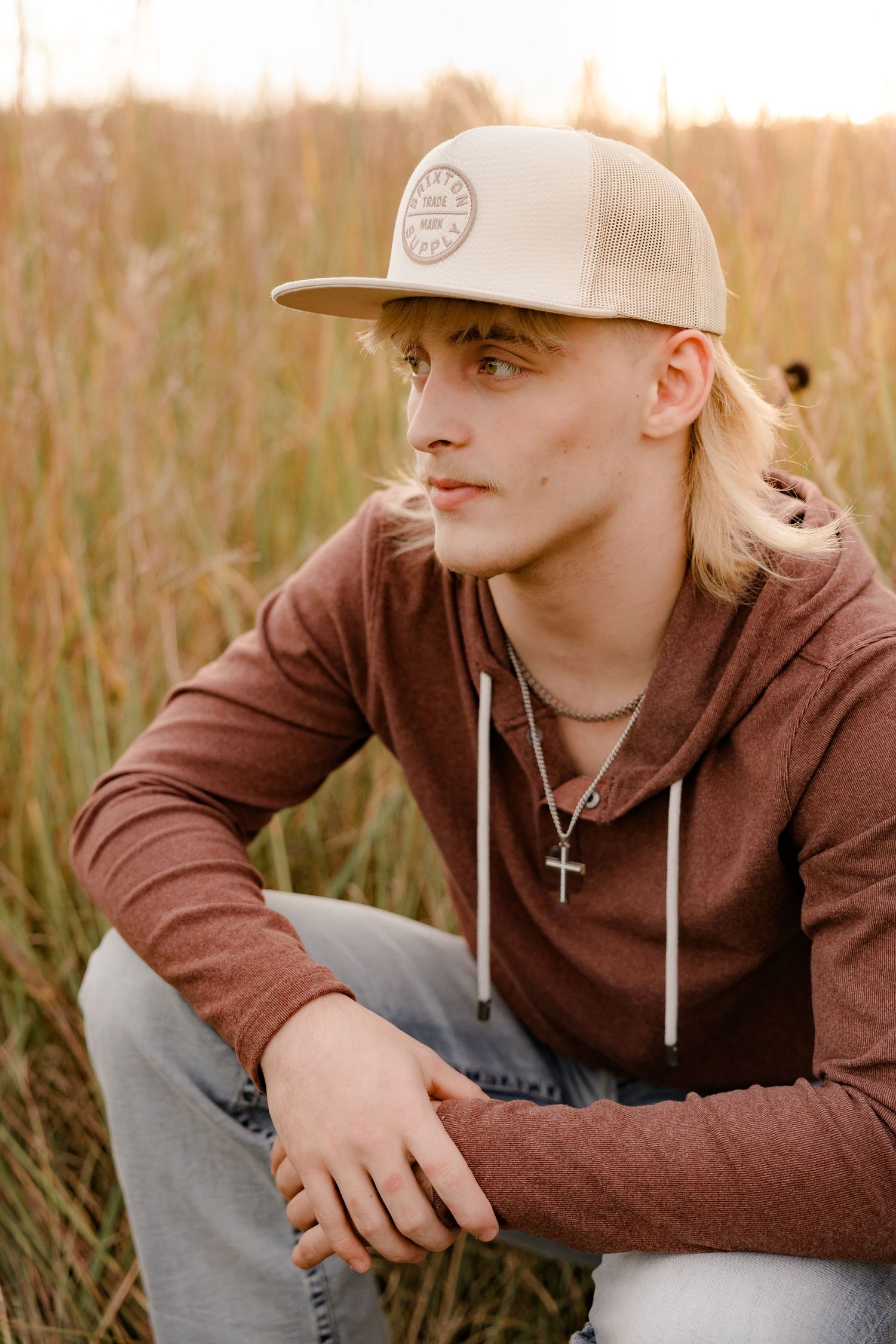 High school senior in brown hoodie and baseball cap sitting thoughtfully in tall grass at sunset.