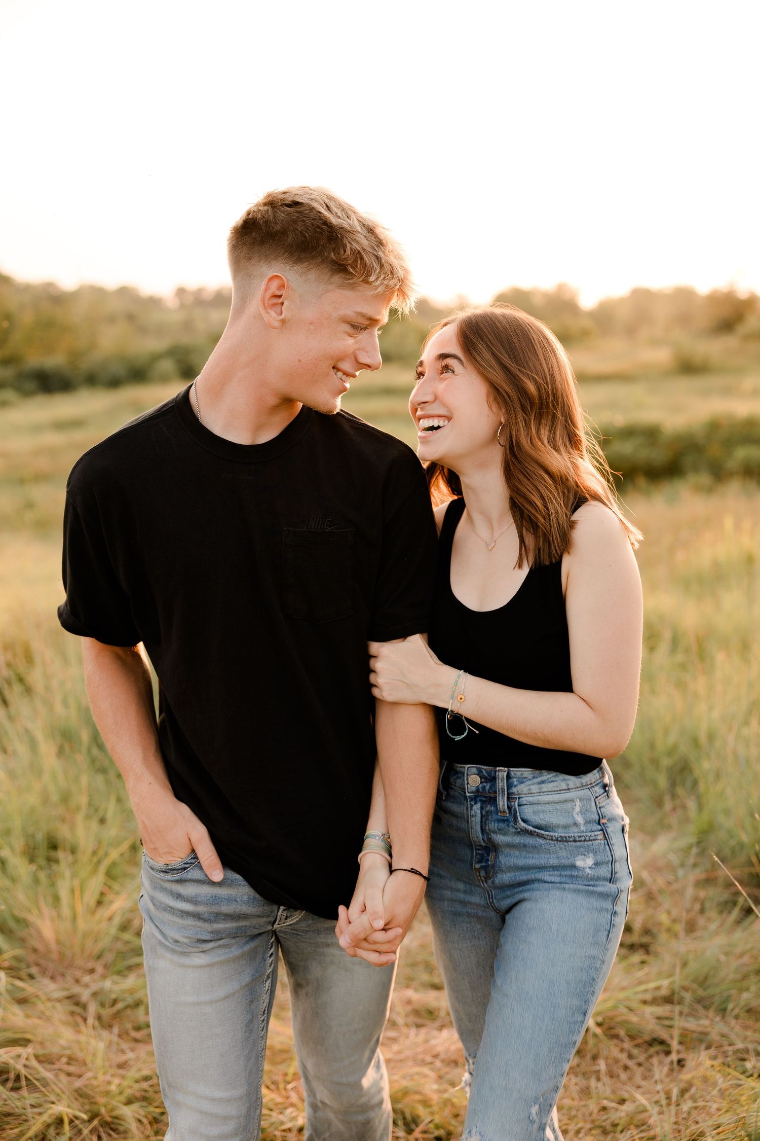 Young couple in matching black shirts and jeans holding hands and laughing together in a golden sunlit field.