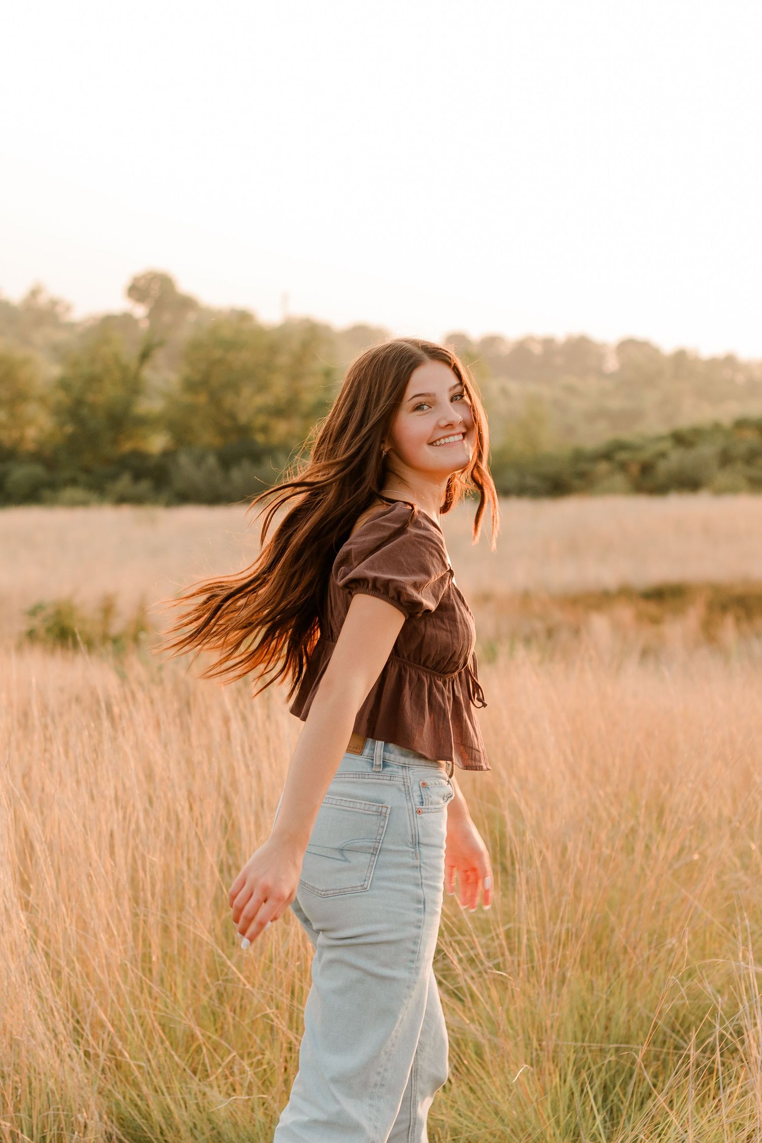 High school senior in brown top and light jeans standing in golden field at sunset with flowing hair and warm lighting.