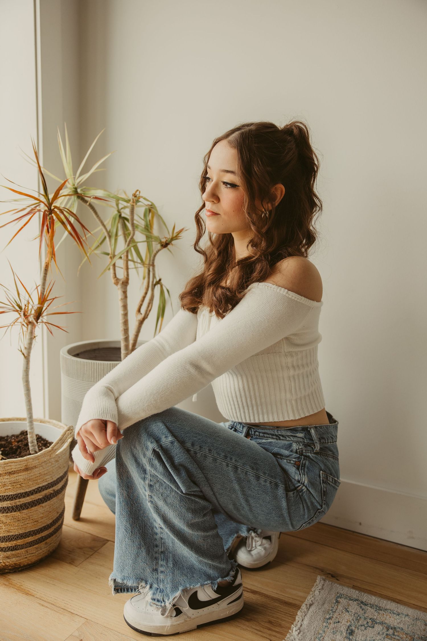 A high school senior in a white cropped sweater and jeans crouches near a potted plant by a window.