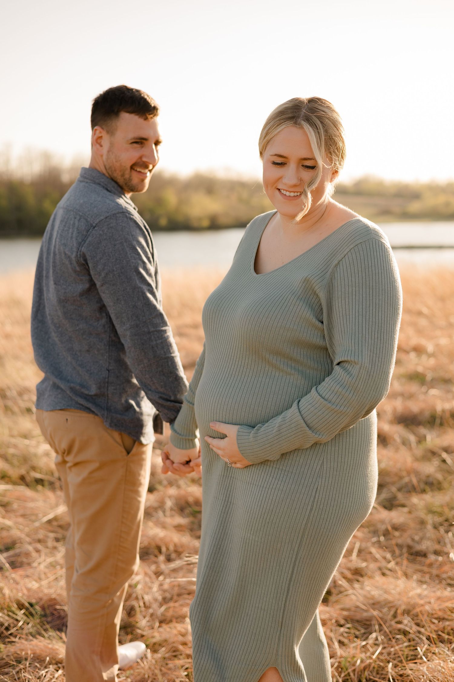 A couple in warm sweaters share a tender moment at sunset by a lake during their maternity photoshoot.