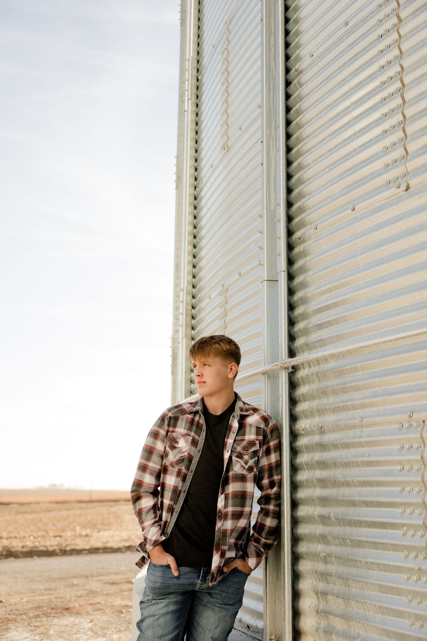 A high school senior in a plaid flannel shirt and jeans leans against a metal grain silo in a rural setting at sunset.