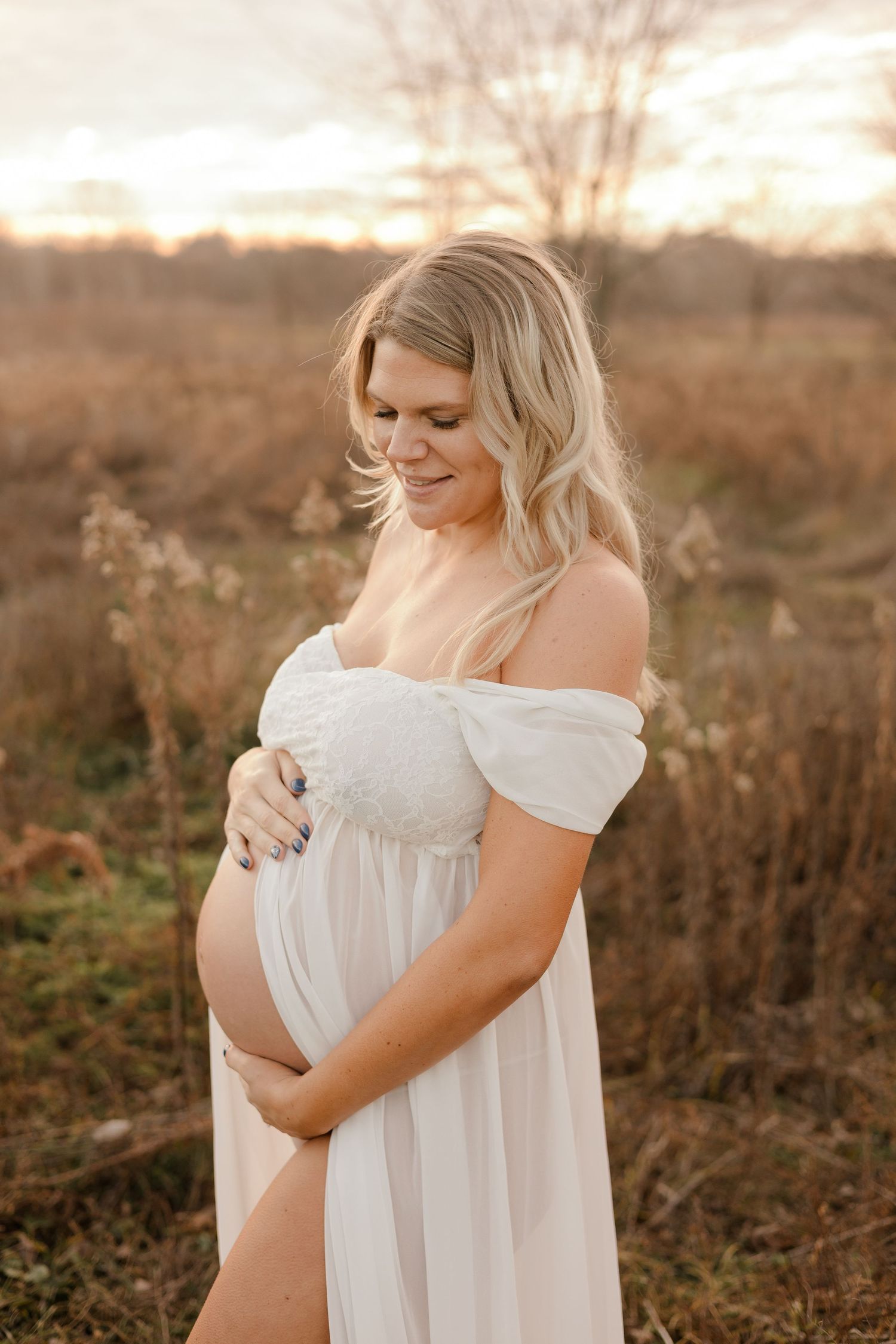 A pregnant person in a flowing white off-shoulder dress poses in a golden field at sunset.