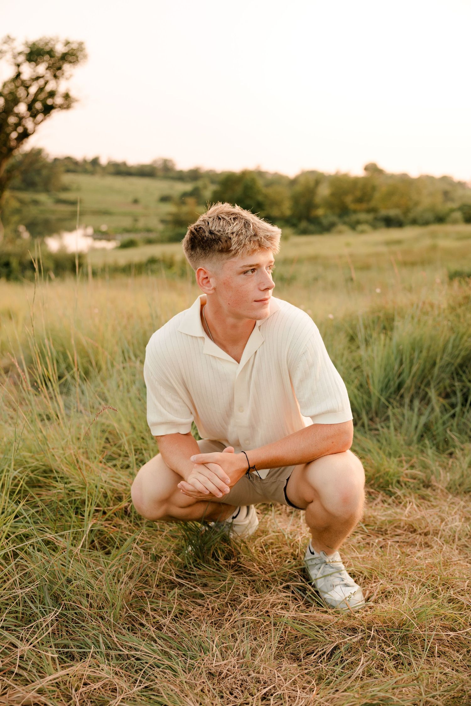 High school senior in white polo shirt crouches in tall grass field during golden hour sunset in rural countryside.