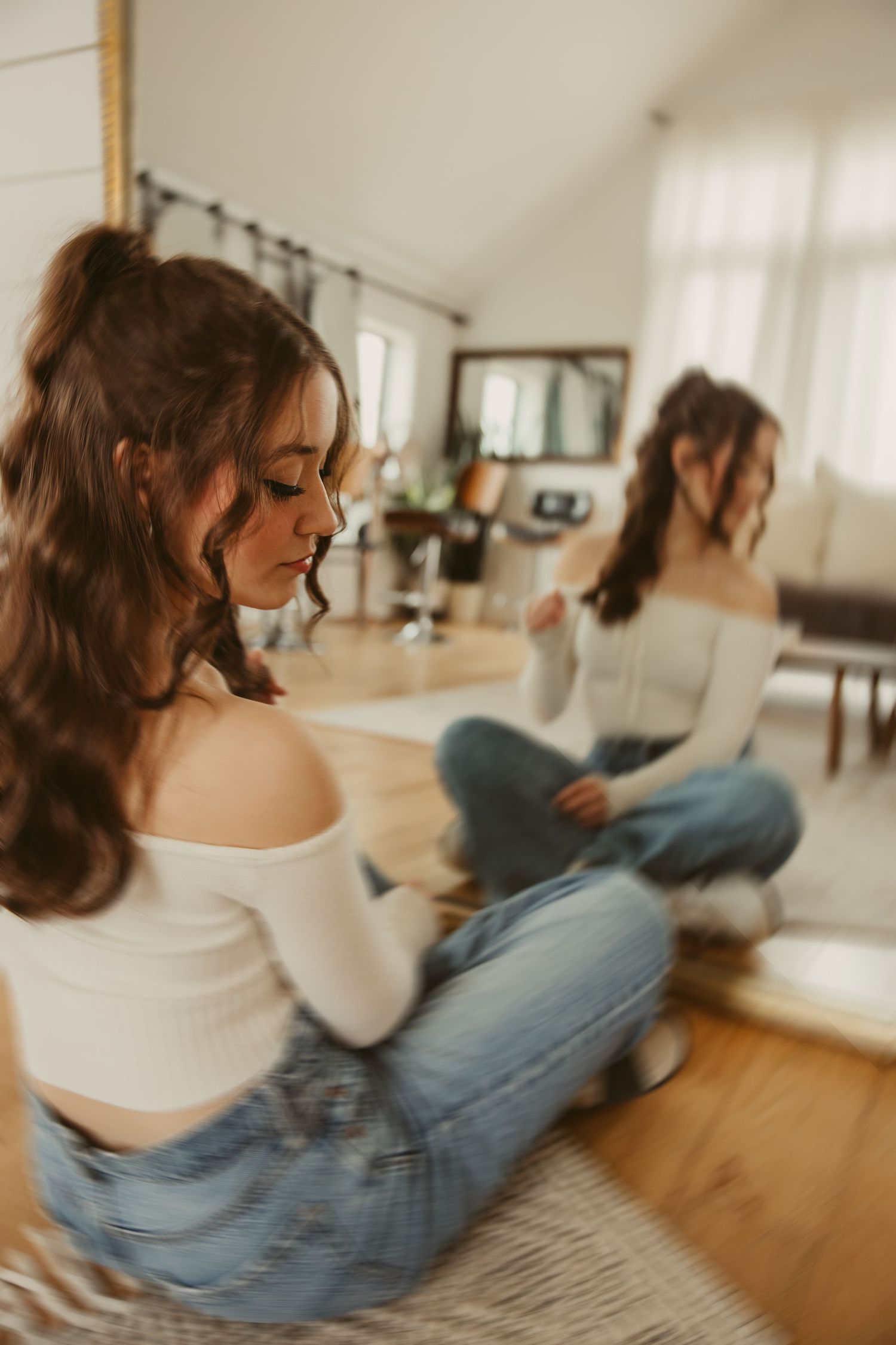 High school senior in white off-shoulder top and jeans sits on hardwood floor beside mirror reflection in bright indoor space.