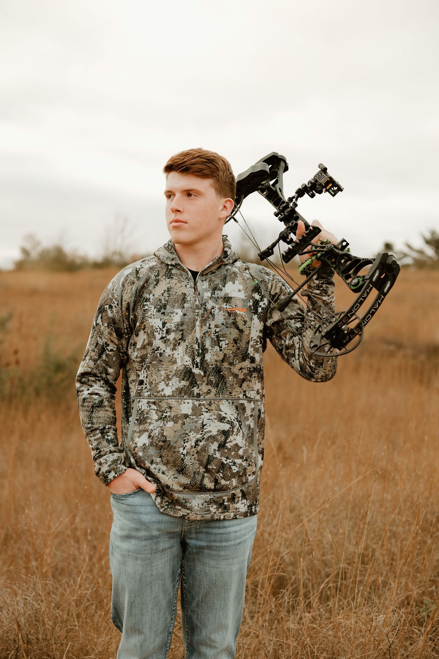 High school senior in camouflage hoodie and jeans holding a compound bow in an autumn field during overcast weather.