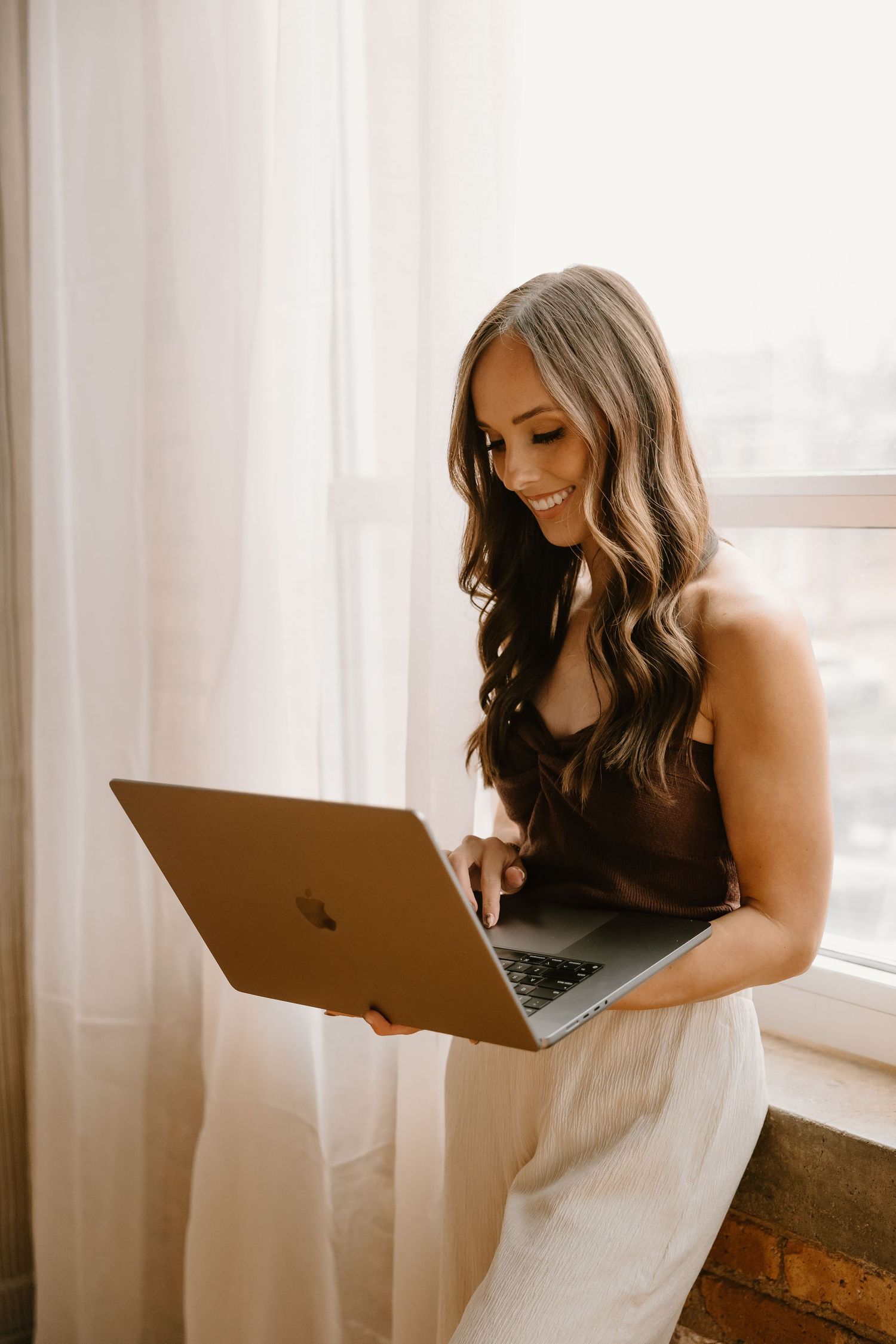 Professional photographer in brown top and white pants sits by window working on MacBook laptop with natural lighting.