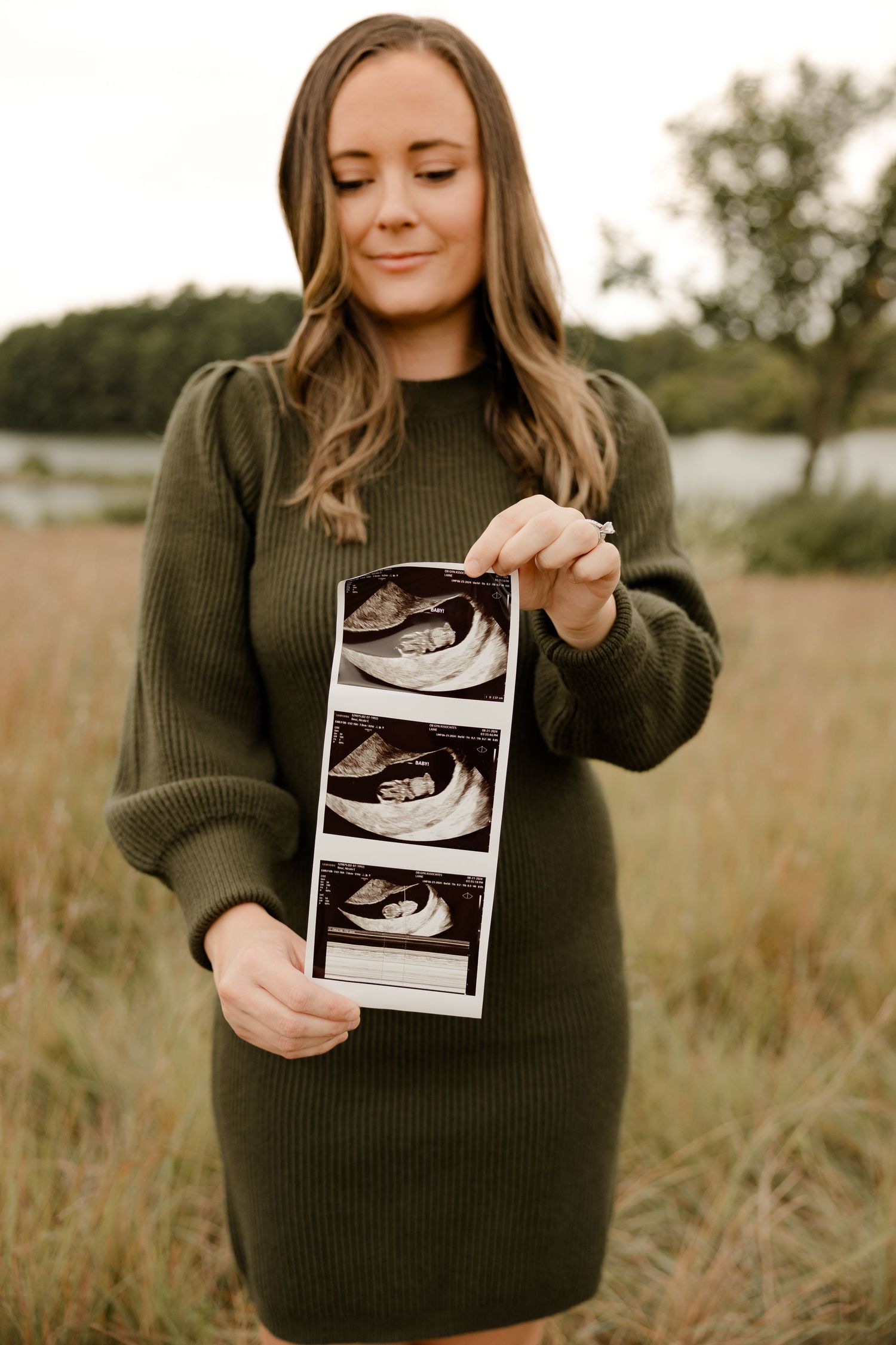 Expecting mom in dark green sweater dress holds ultrasound photos while standing in a grassy field during autumn.