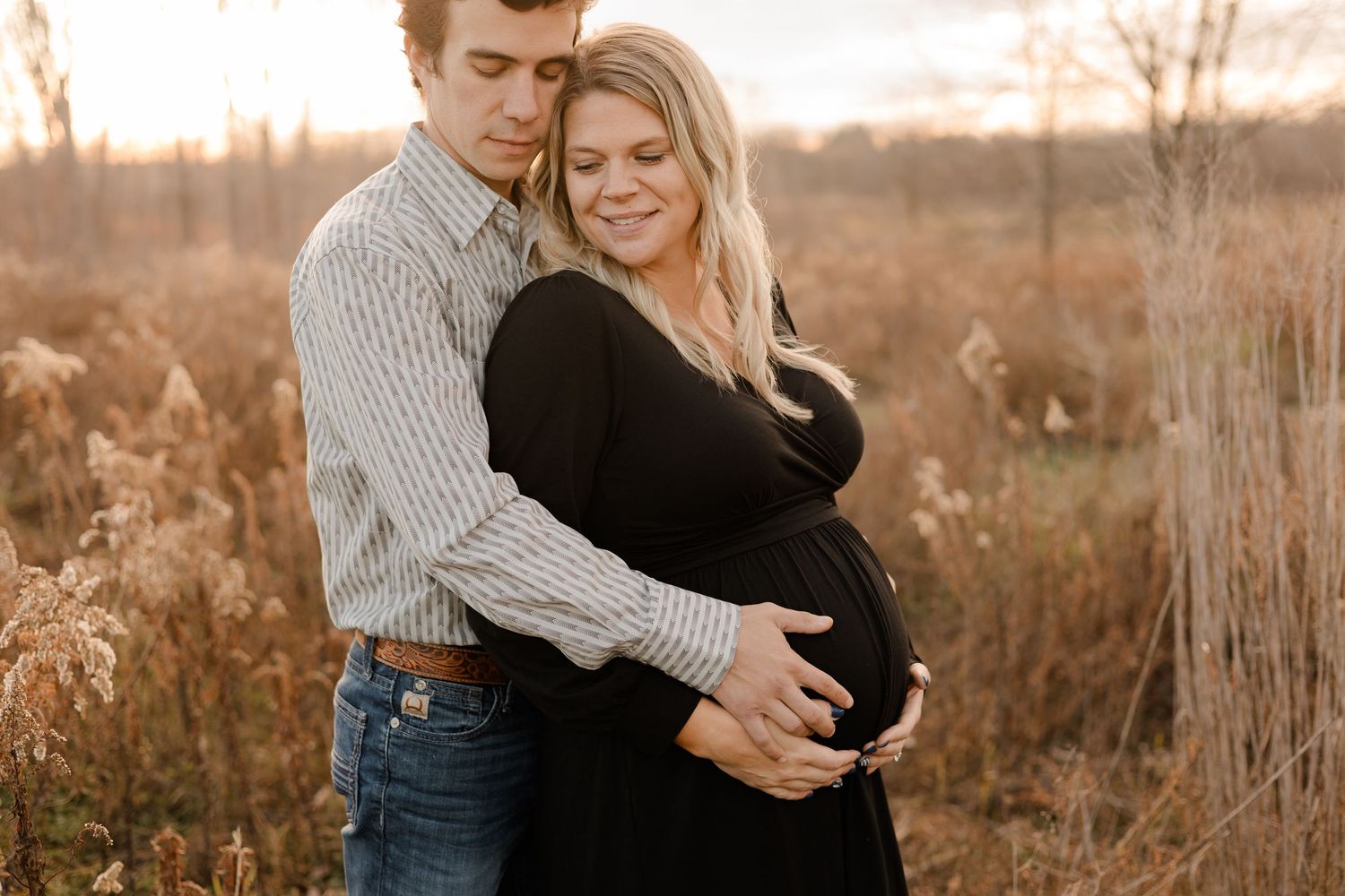 A couple embraces in a golden field during sunset, with tall grass and warm autumn colors surrounding them.