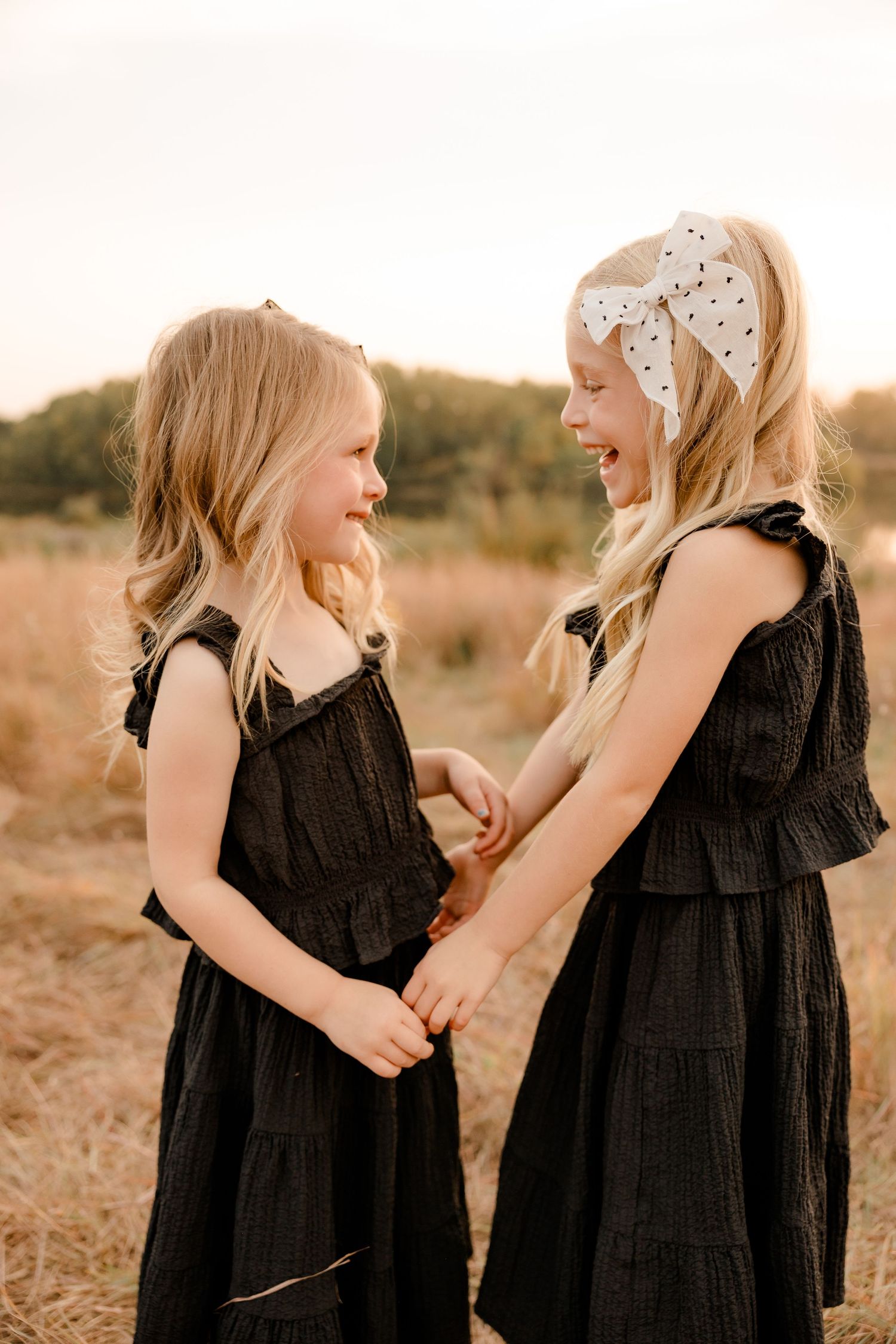 Two sisters in matching black dresses share a joyful moment together in a golden field at sunset.