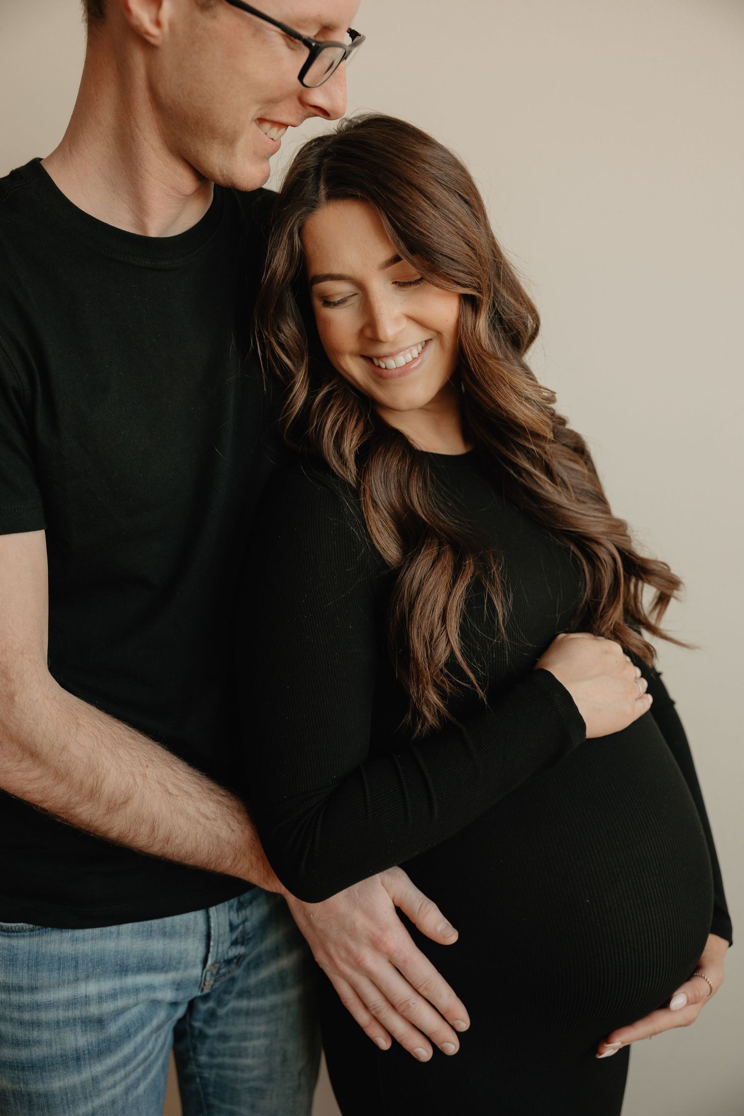 A couple in matching black tops pose for an intimate maternity photo while smiling against a neutral backdrop.