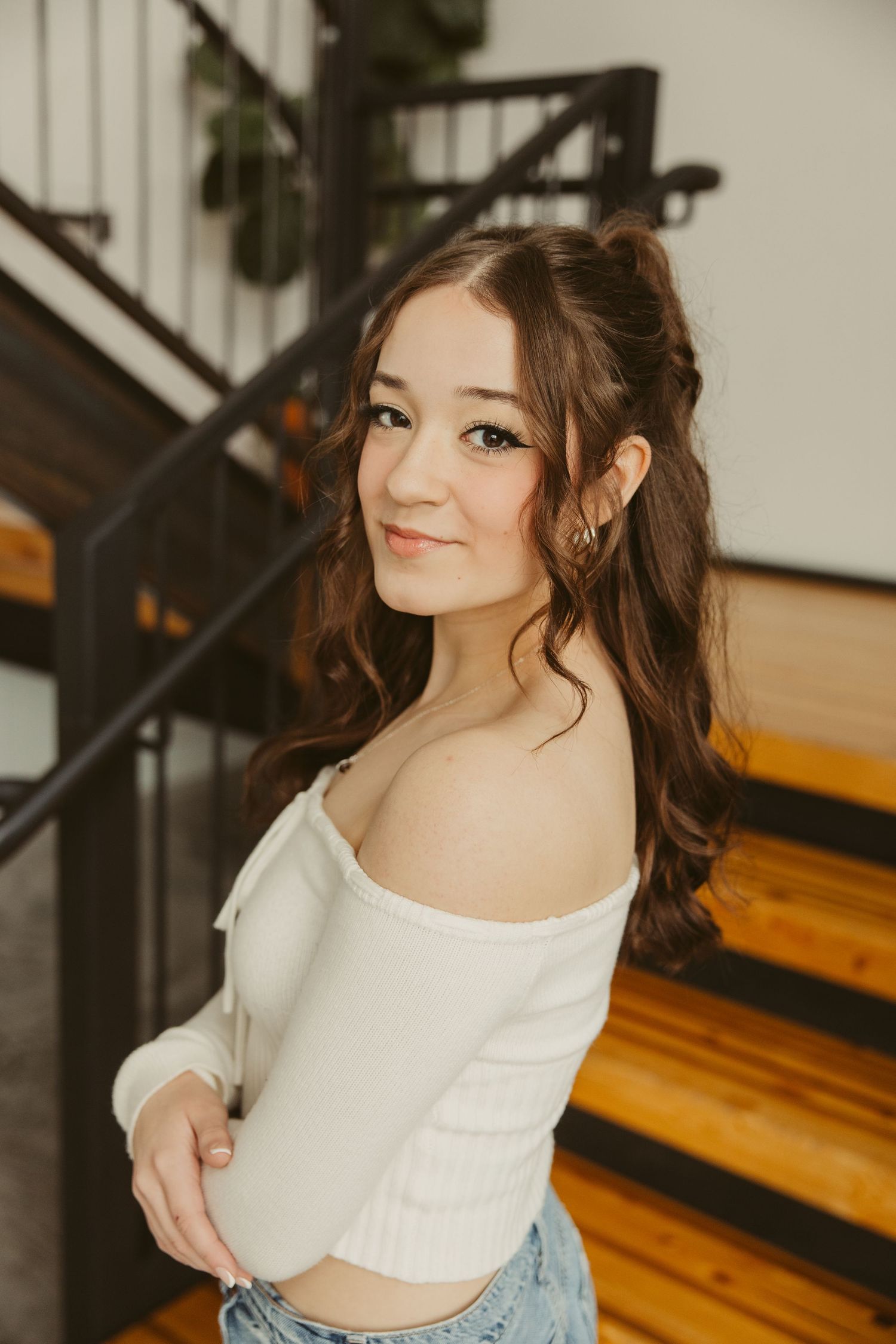 A stylish high school senior in a white crop top and jeans poses on wooden stairs with a casual, confident smile.