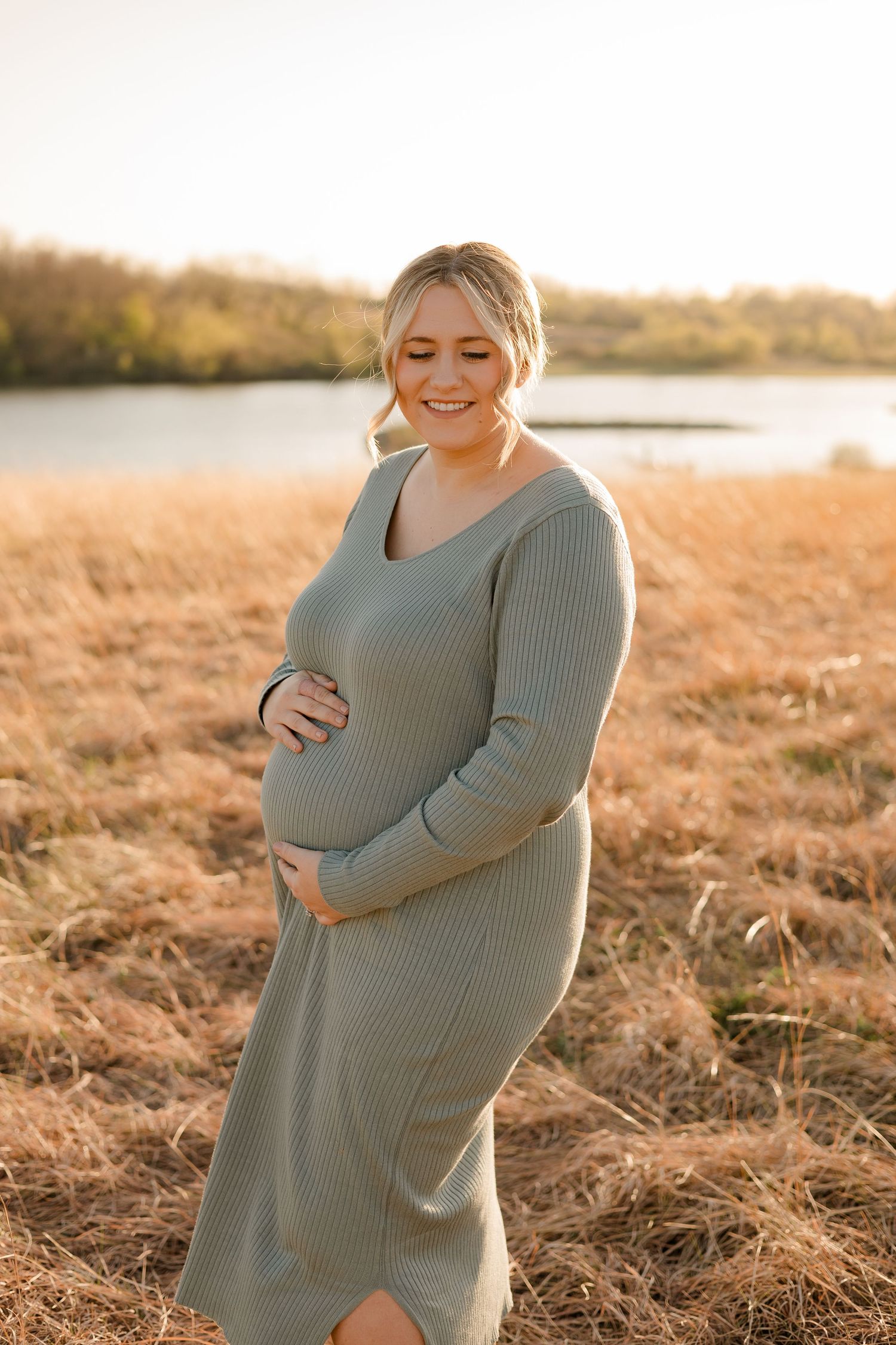 A pregnant woman in a gray sweater dress stands in a golden field at sunset, smiling and cradling her baby bump.