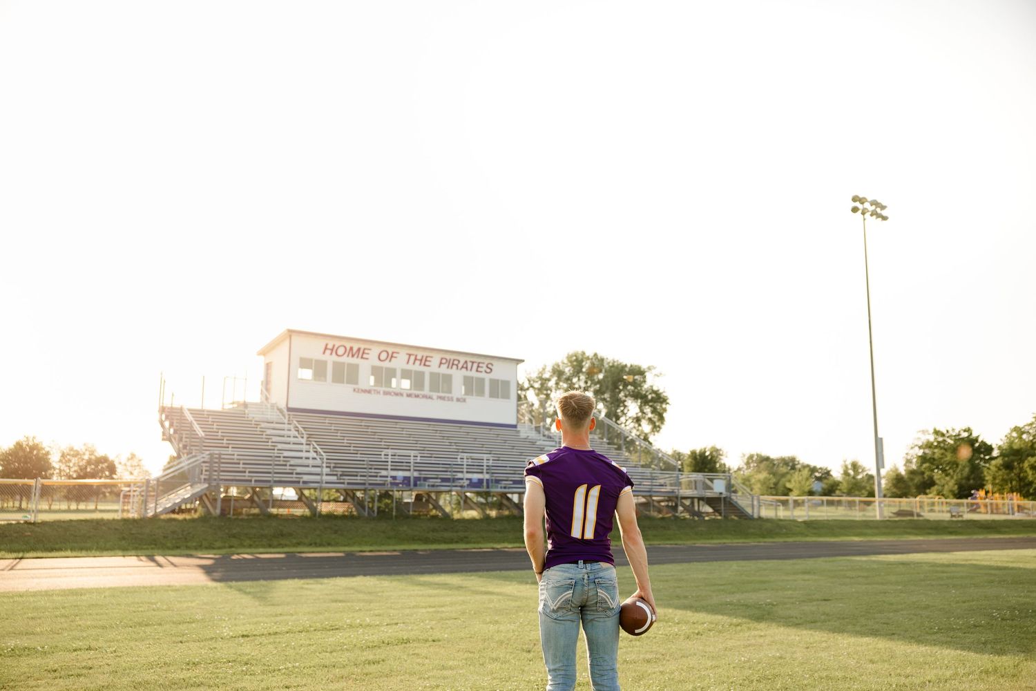 Football player in jersey number 11 stands on the field at sunset with stadium bleachers in the background.