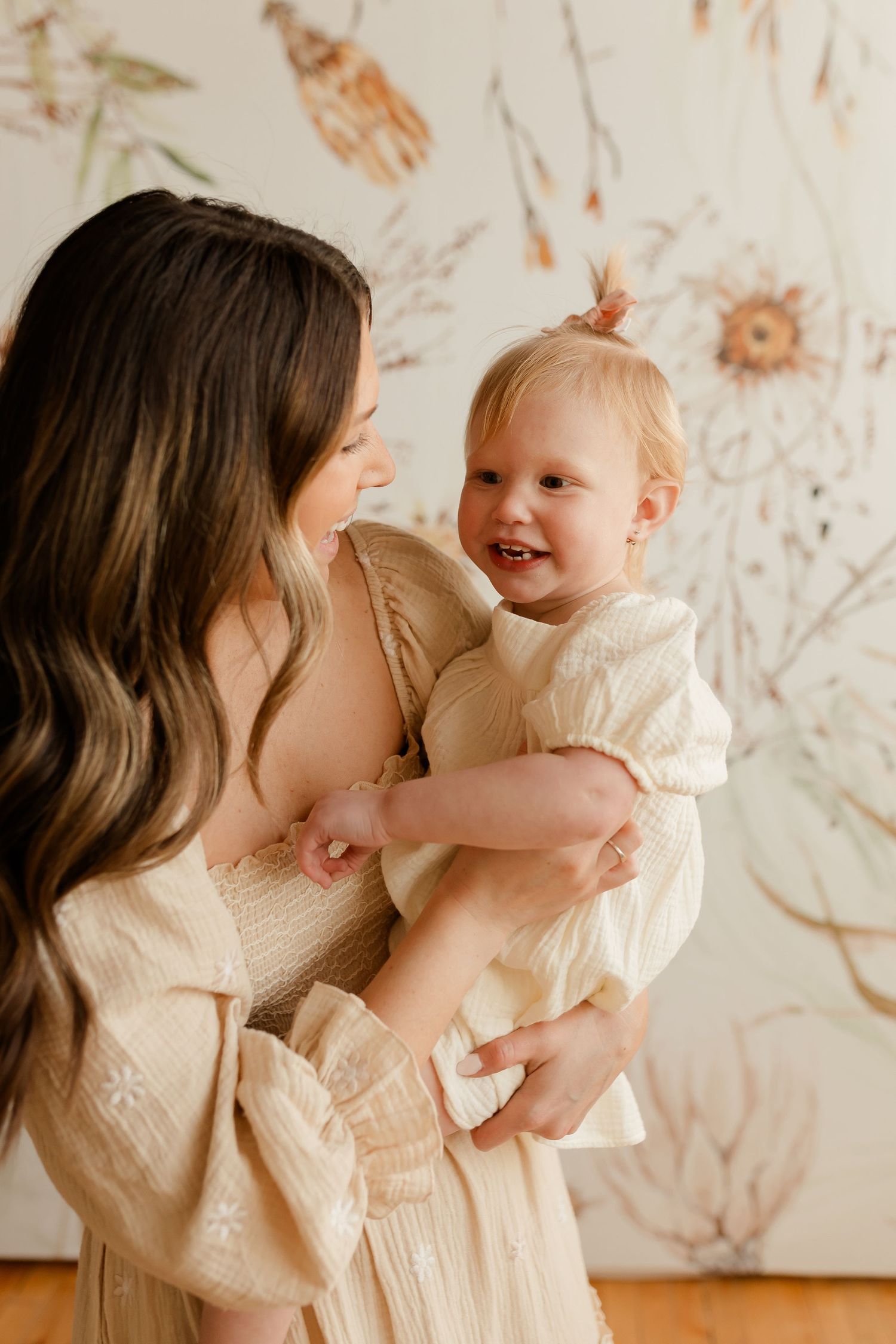 A mother and baby share a joyful moment together while wearing cream-colored dresses against a floral wallpaper background.
