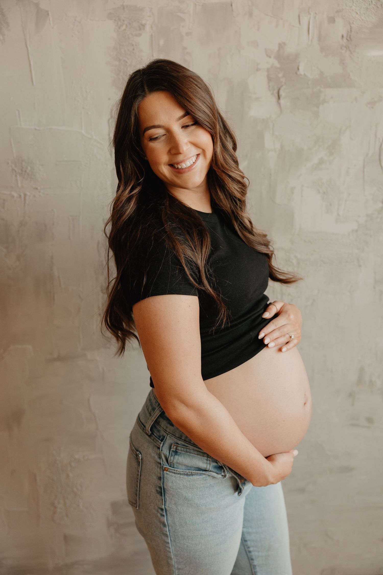 A pregnant woman in a black t-shirt and jeans poses happily against a textured gray wall.