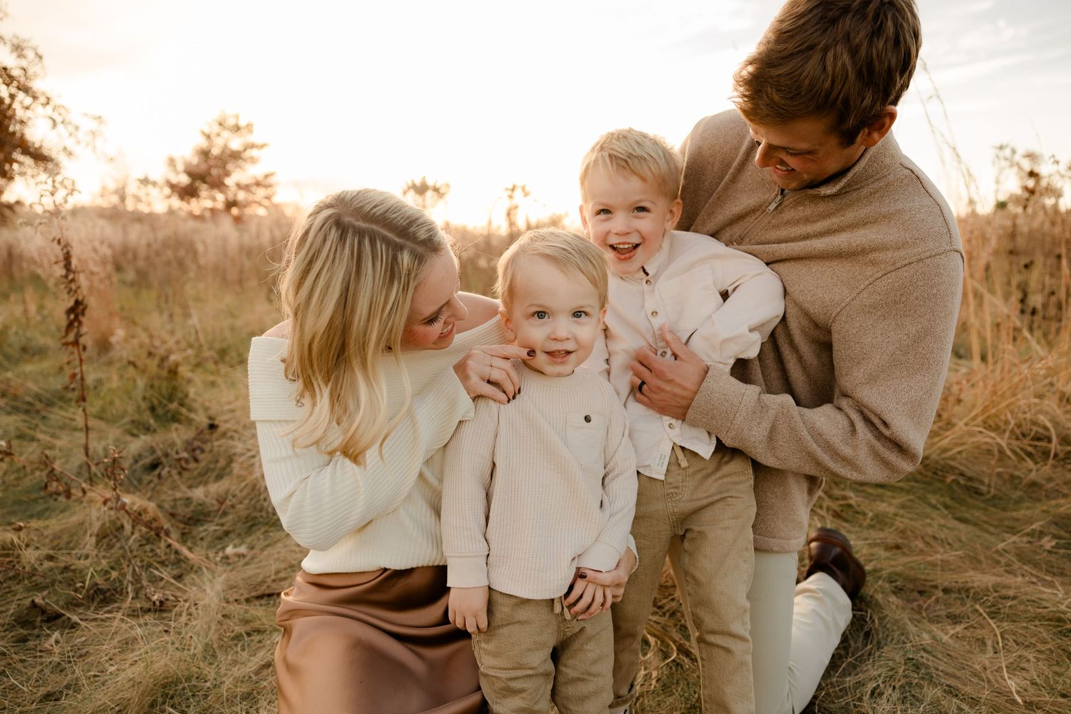 A family sits together in a golden field during sunset while wearing cozy neutral-colored clothing.