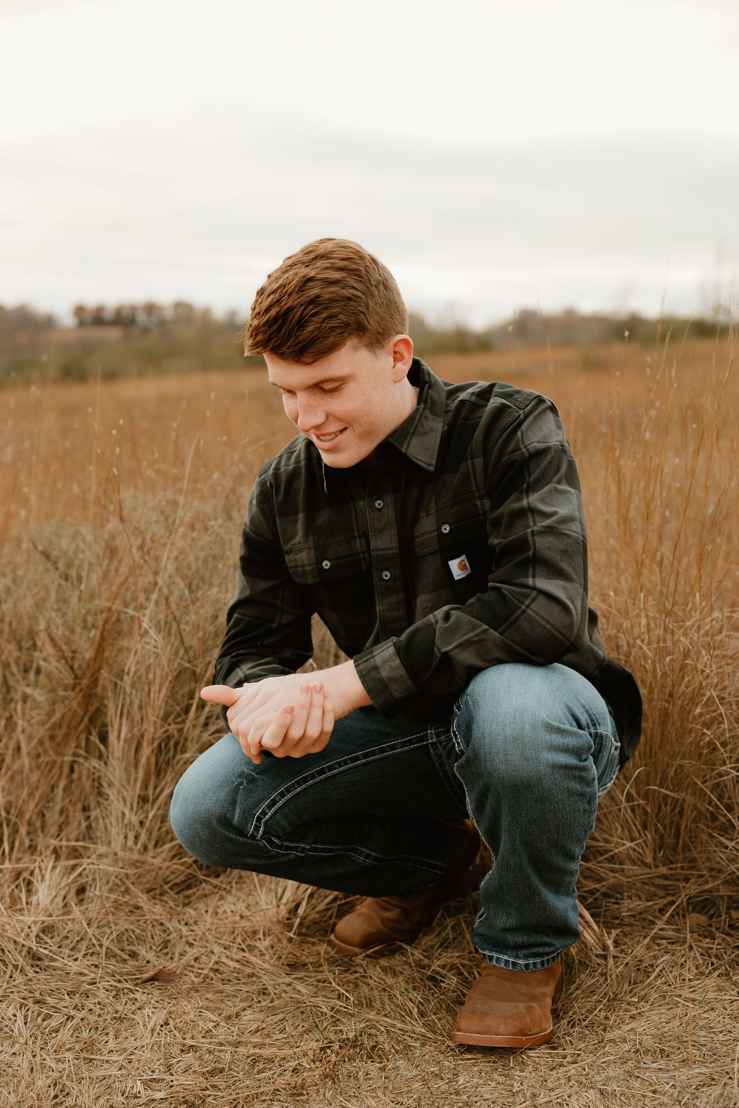 High school senior in dark jacket and jeans crouches in tall autumn grass field during golden hour.