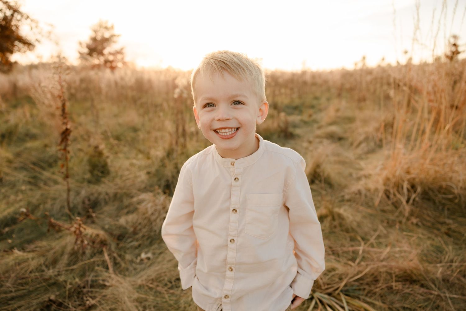 A child wearing a cream colored button-up cardigan stands in a golden field at sunset smiling warmly.