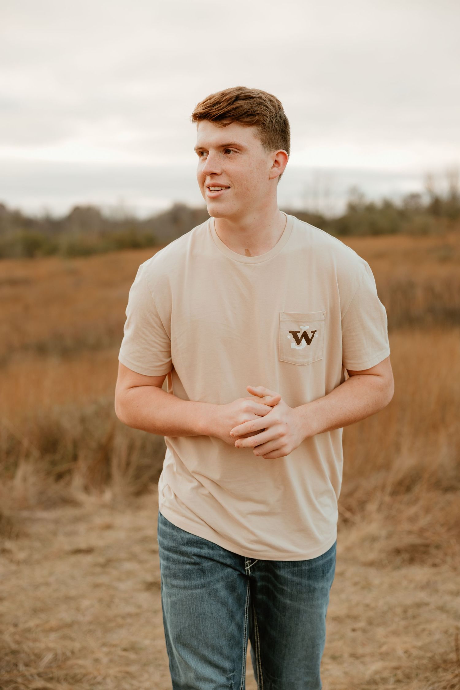 A high school senior wearing a beige t-shirt with a W logo and jeans stands in a golden field during sunset.