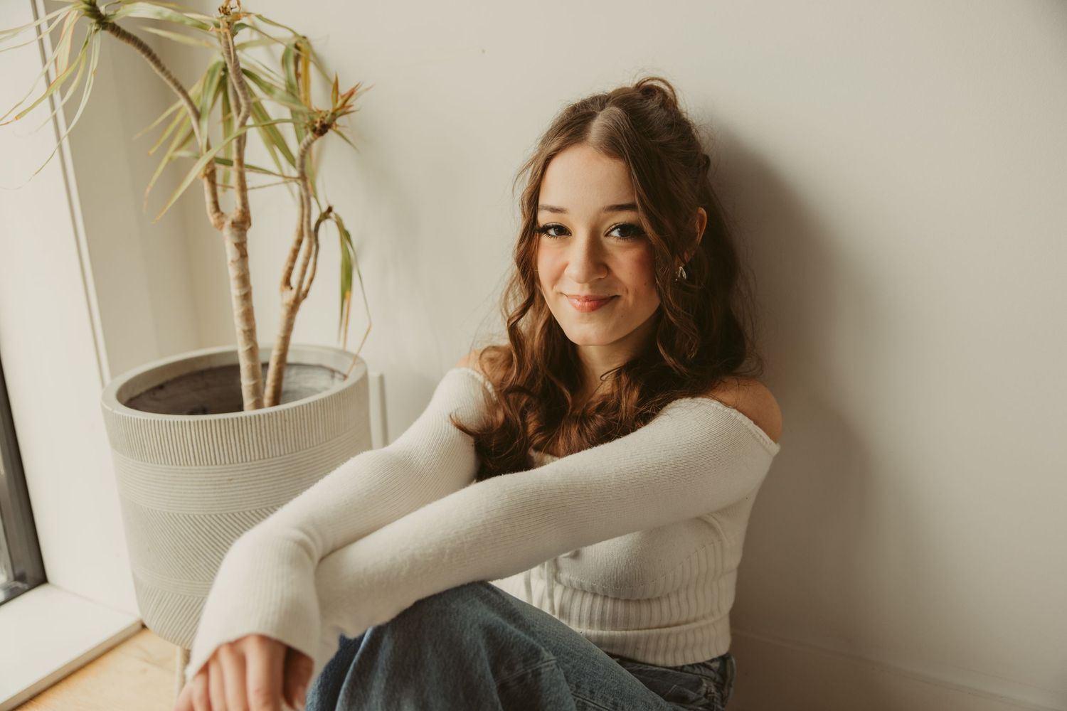 High school senior in white sweater sitting casually by window with potted bamboo plant in natural lighting.