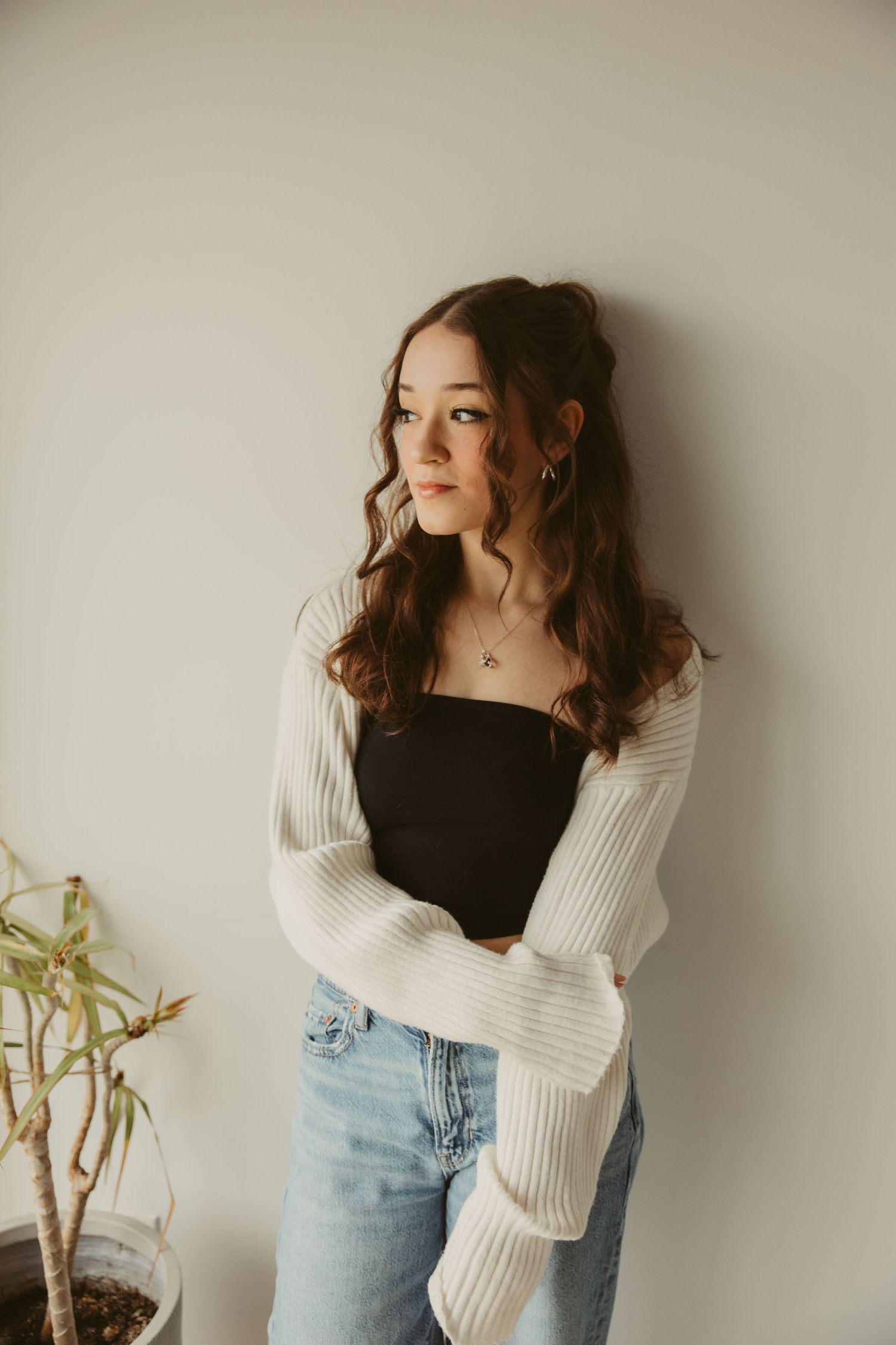 A high school senior in casual attire with a white knit cardigan and jeans poses against a neutral wall near a potted plant.