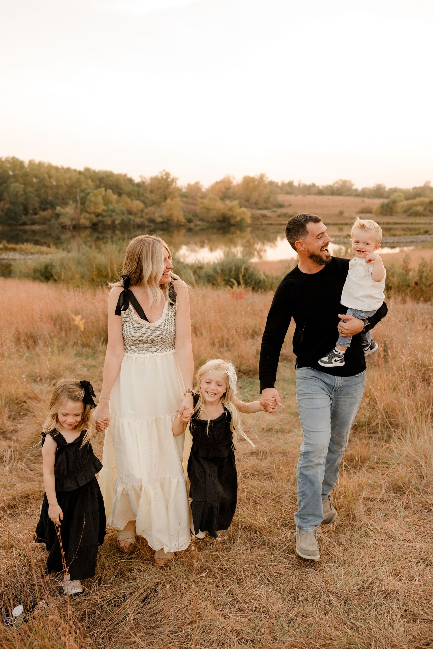 Family walking together in golden field at sunset near a lake wearing coordinated black and white outfits.