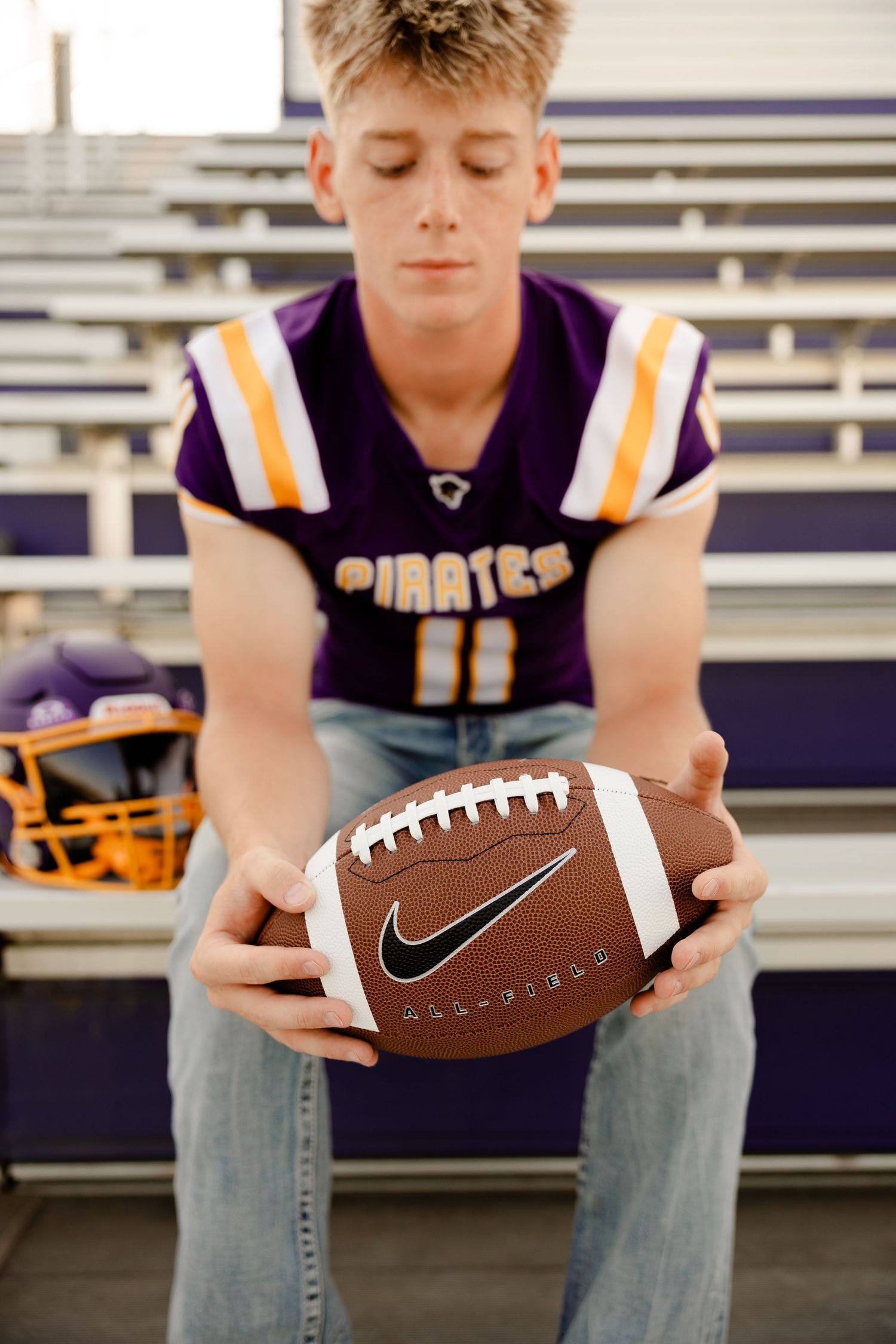 A high school football player wearing purple Pirates jersey holds a Nike football while sitting on bleachers.